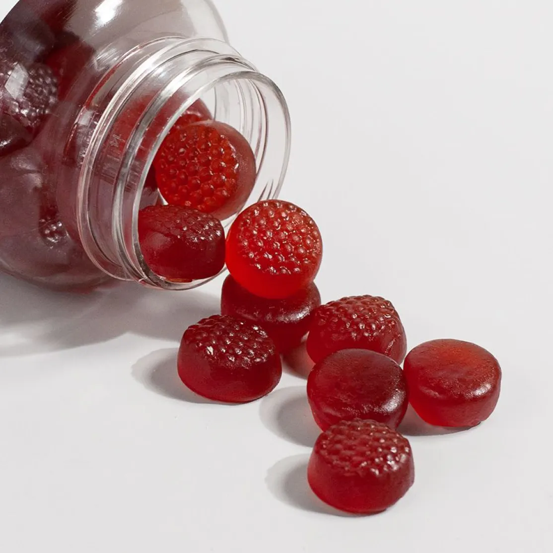 Red sleep gummies spilling out of a clear plastic jar onto a white surface.