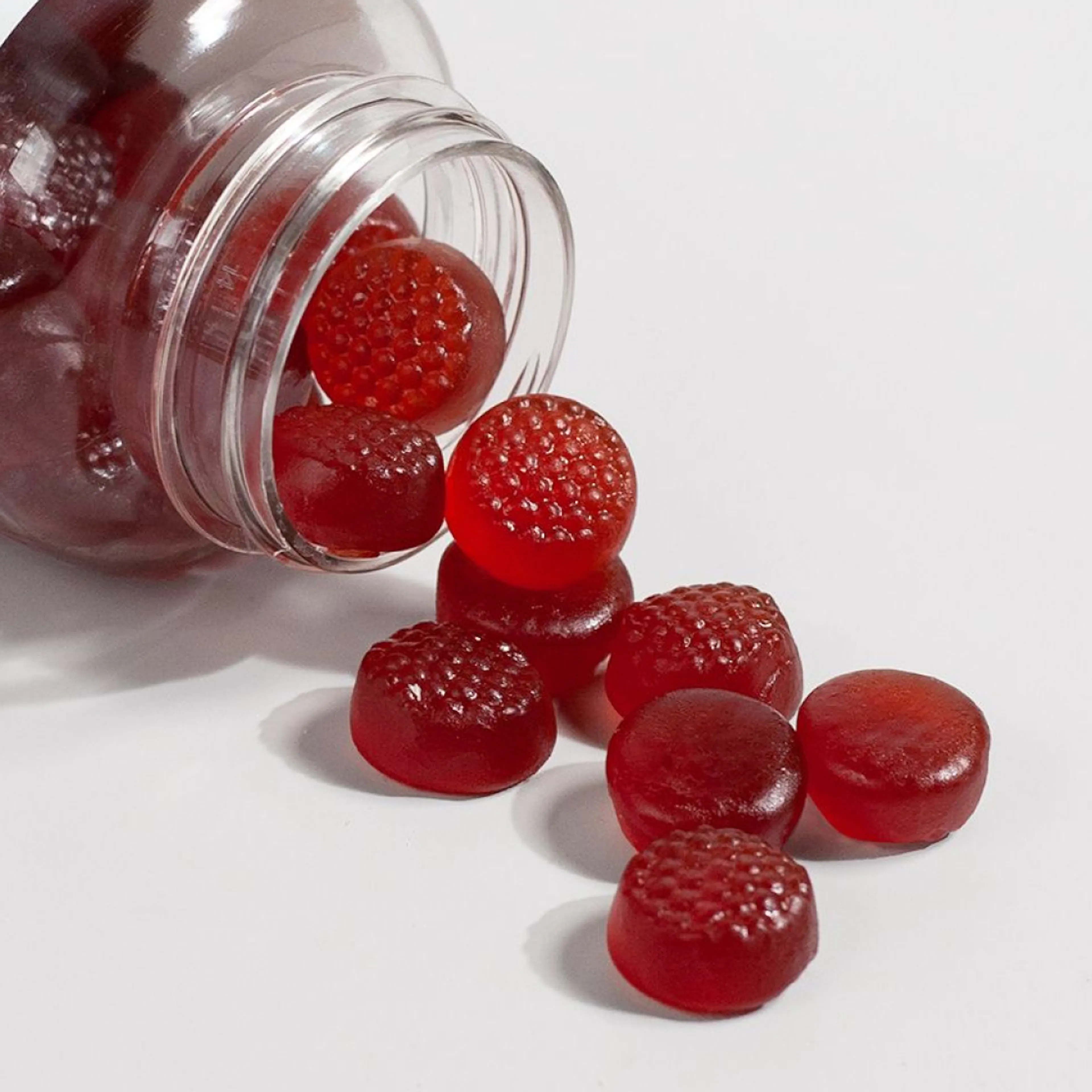 Red sleep gummies spilling out of a clear plastic jar onto a white surface.