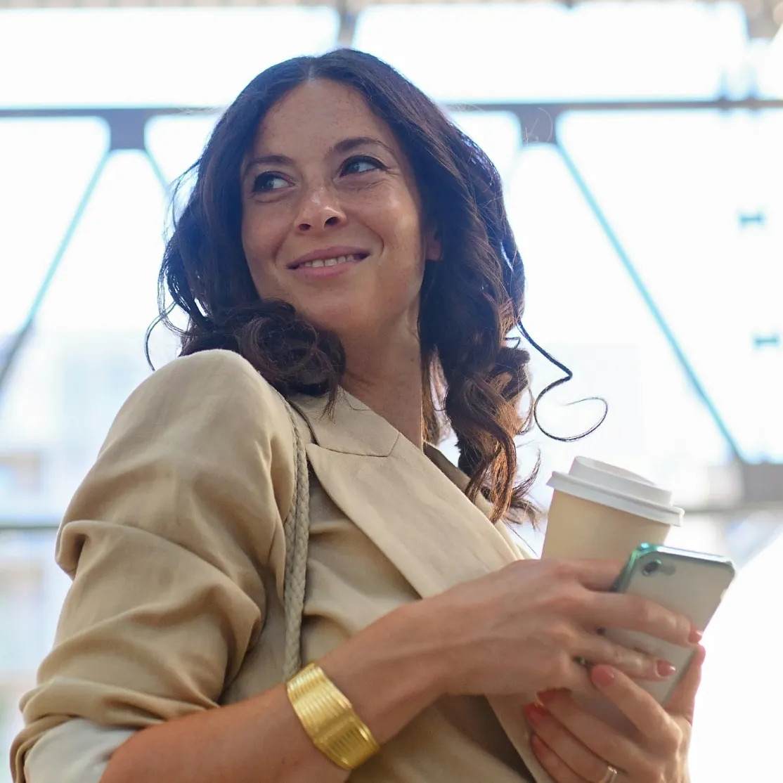 A woman in midlife holding a coffee and her phone, smiling and looking over her shoulder.