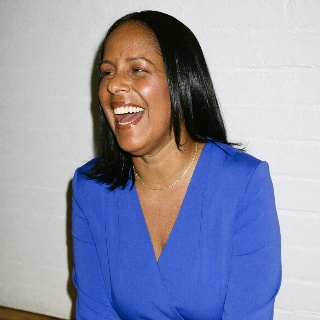 A woman in midlife smiling. She's in a blue shirt and sitting in front of a white wall.
