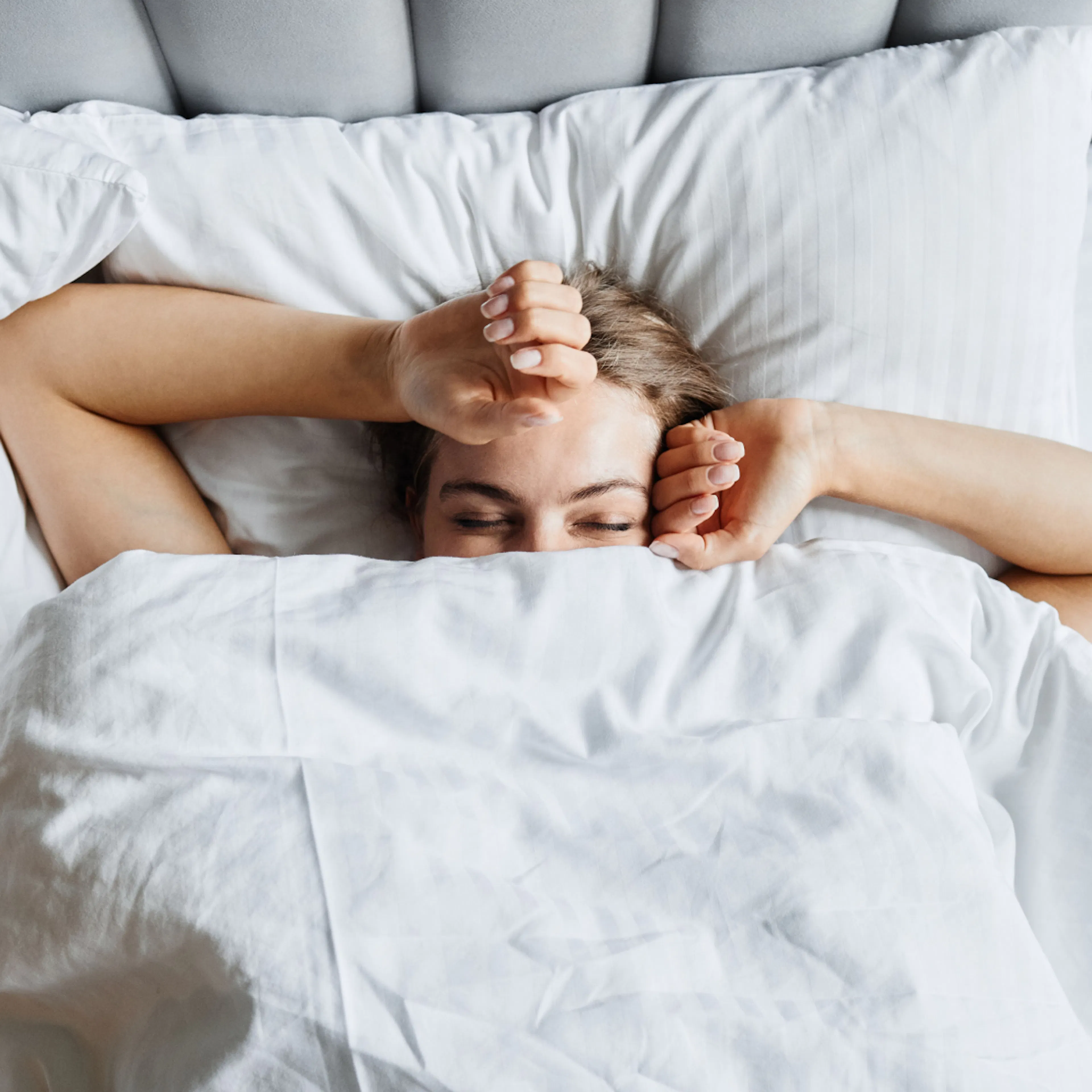 A woman happily waking up. Her face is peeking out from under the covers and her arms are stretched over her head.
