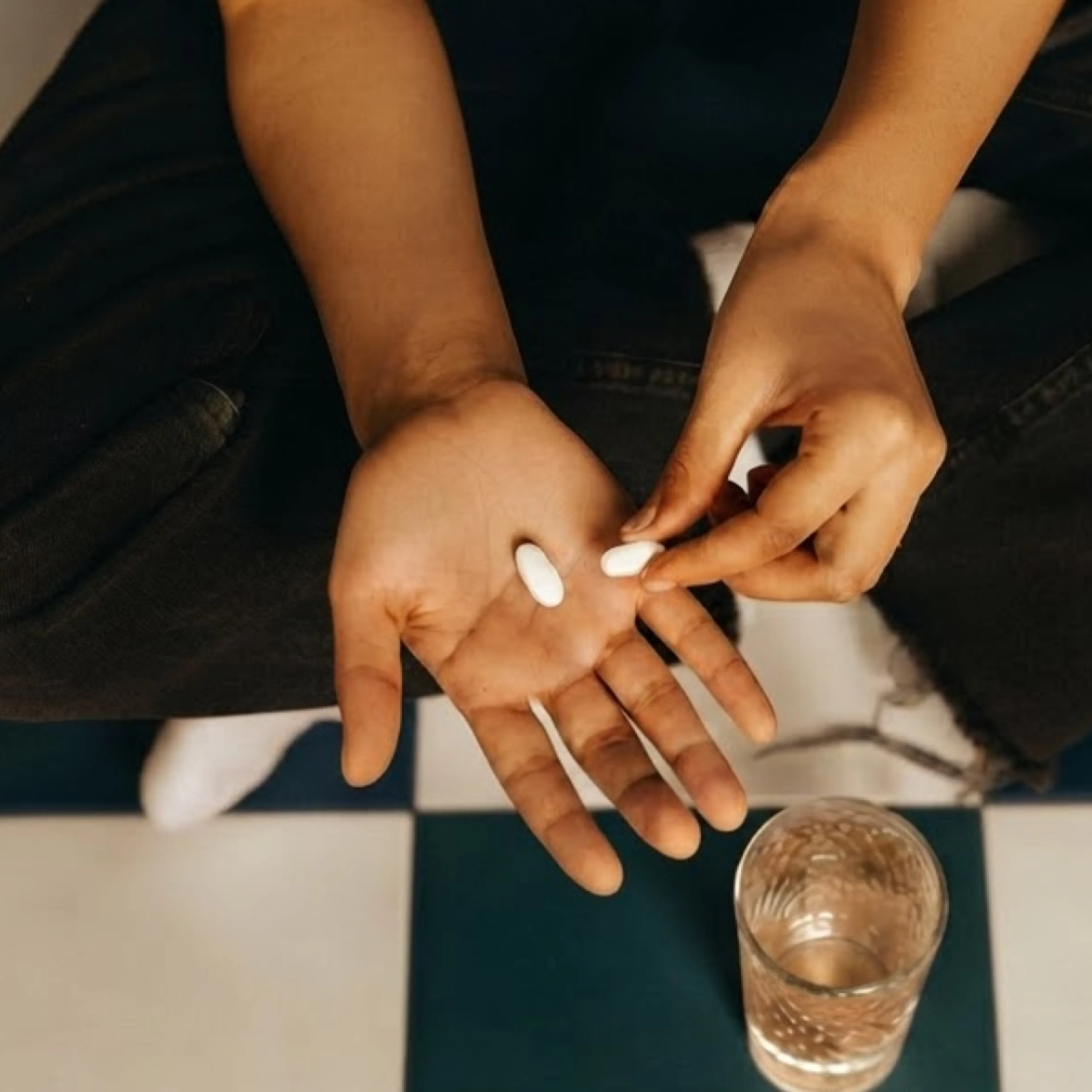 A close-up photo of a woman holding white supplements with a glass of water in the background.