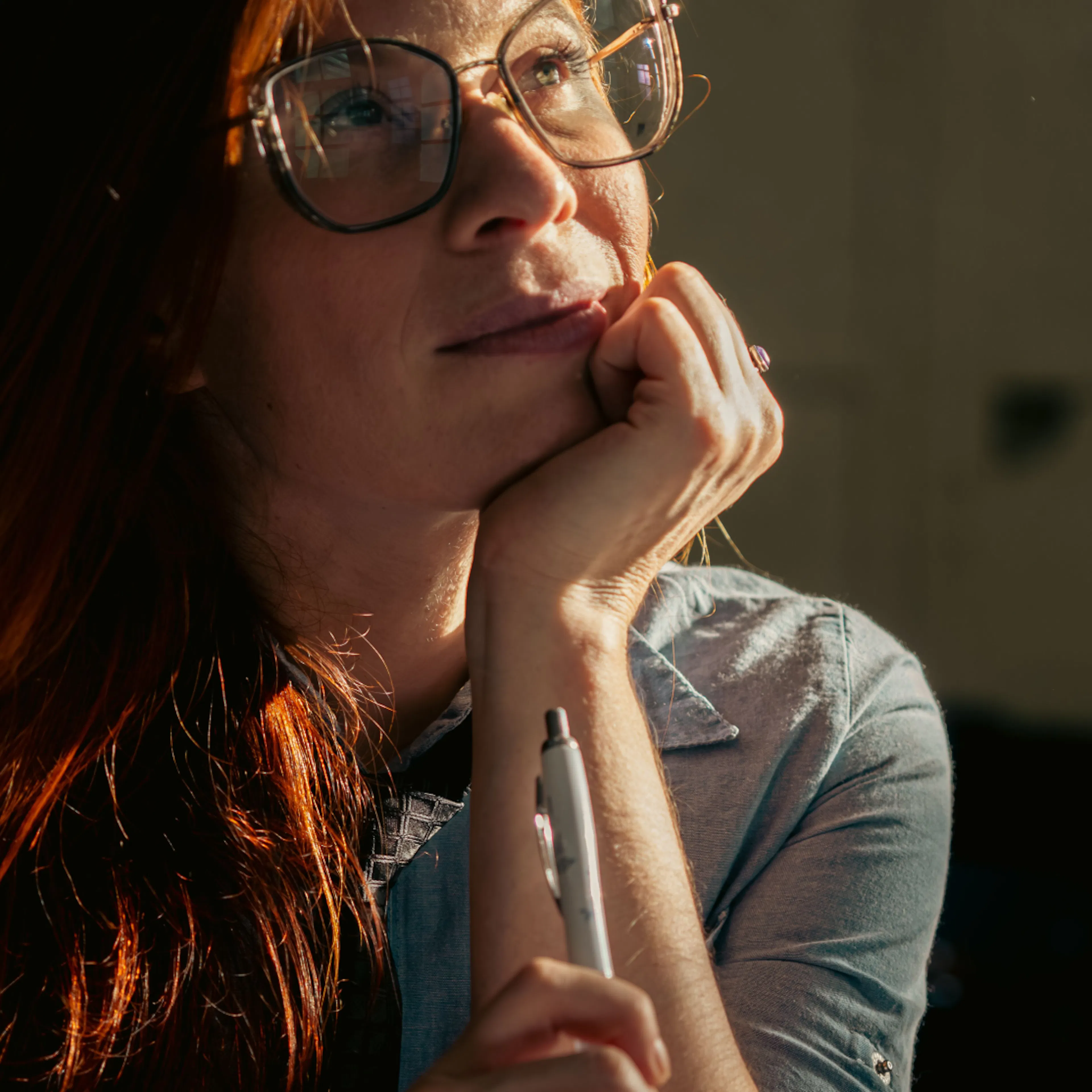 A woman in midlife with her head on her hand, thinking and looking up. She's wearing glasses and has red hair. 