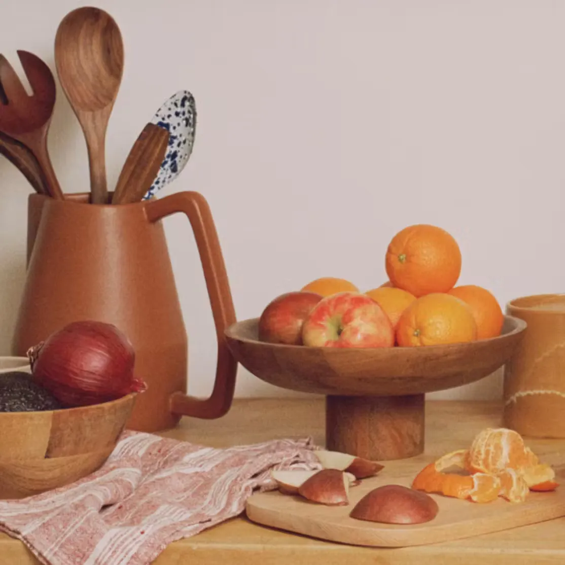A kitchen counter scene featuring a pedestal wooden bowl filled with fresh oranges and apples, a wooden cutting board with sliced fruit, a terracotta pitcher holding wooden spoons, and a red onion in a bowl