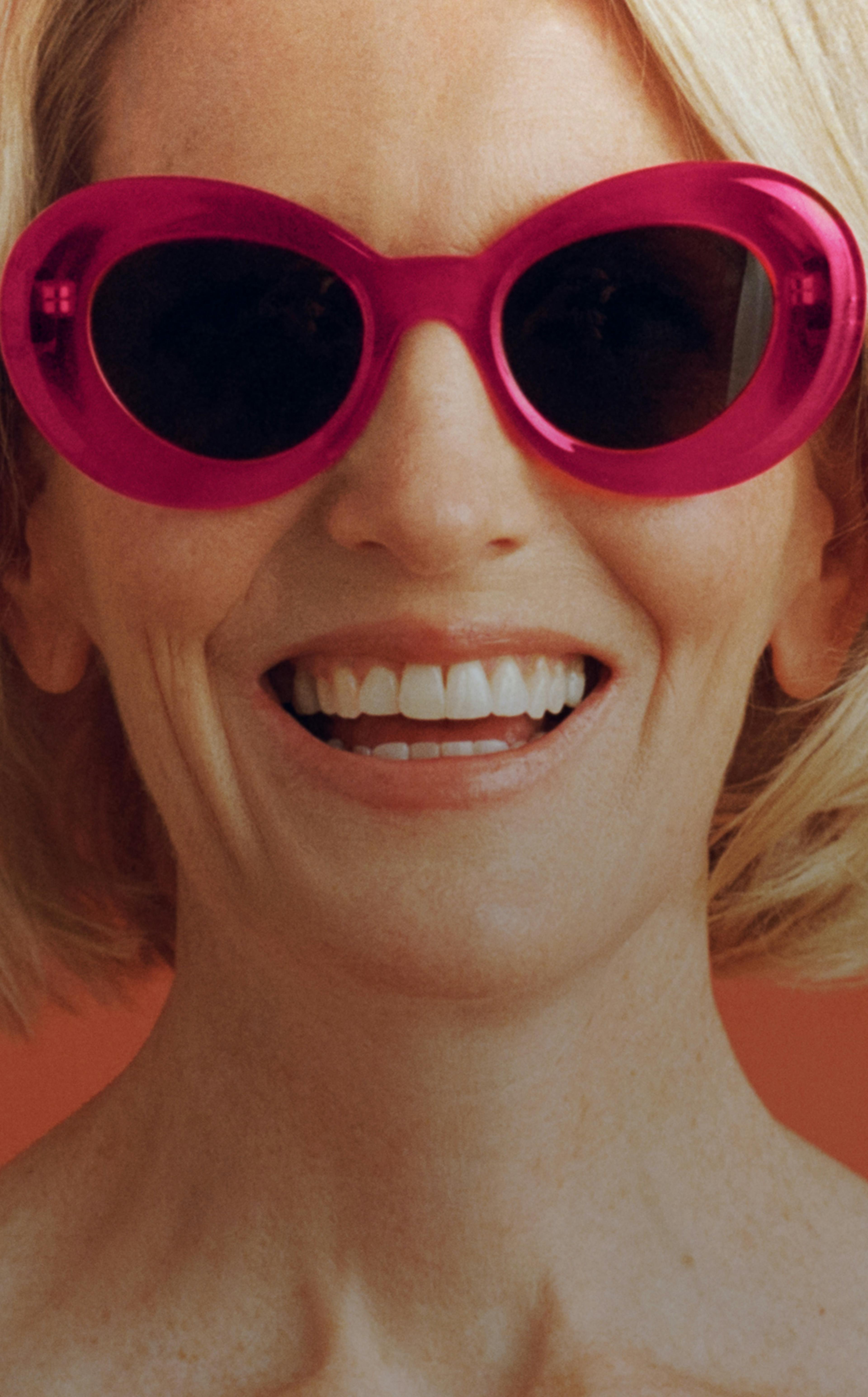 Portrait photograph of woman smiling while wearing hot pink sunglasses