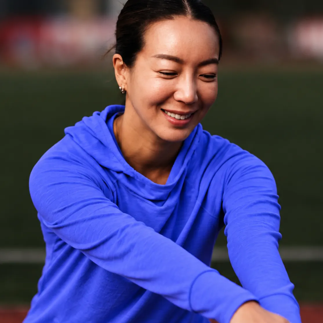 A woman in midlife hanging out outside, smiling and stretching her arms. She's wearing a blue sweatshirt.