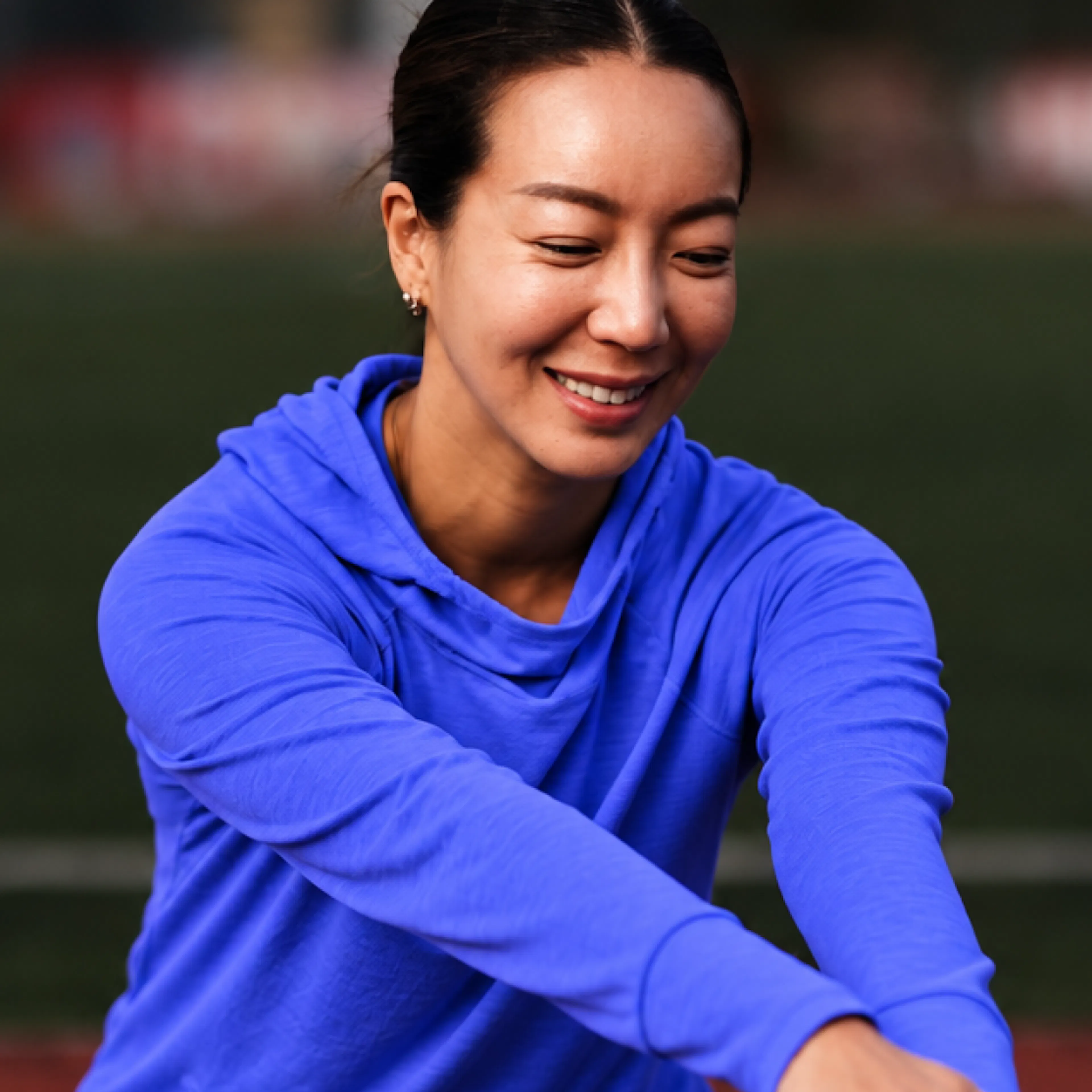 A woman in midlife hanging out outside, smiling and stretching her arms. She's wearing a blue sweatshirt.