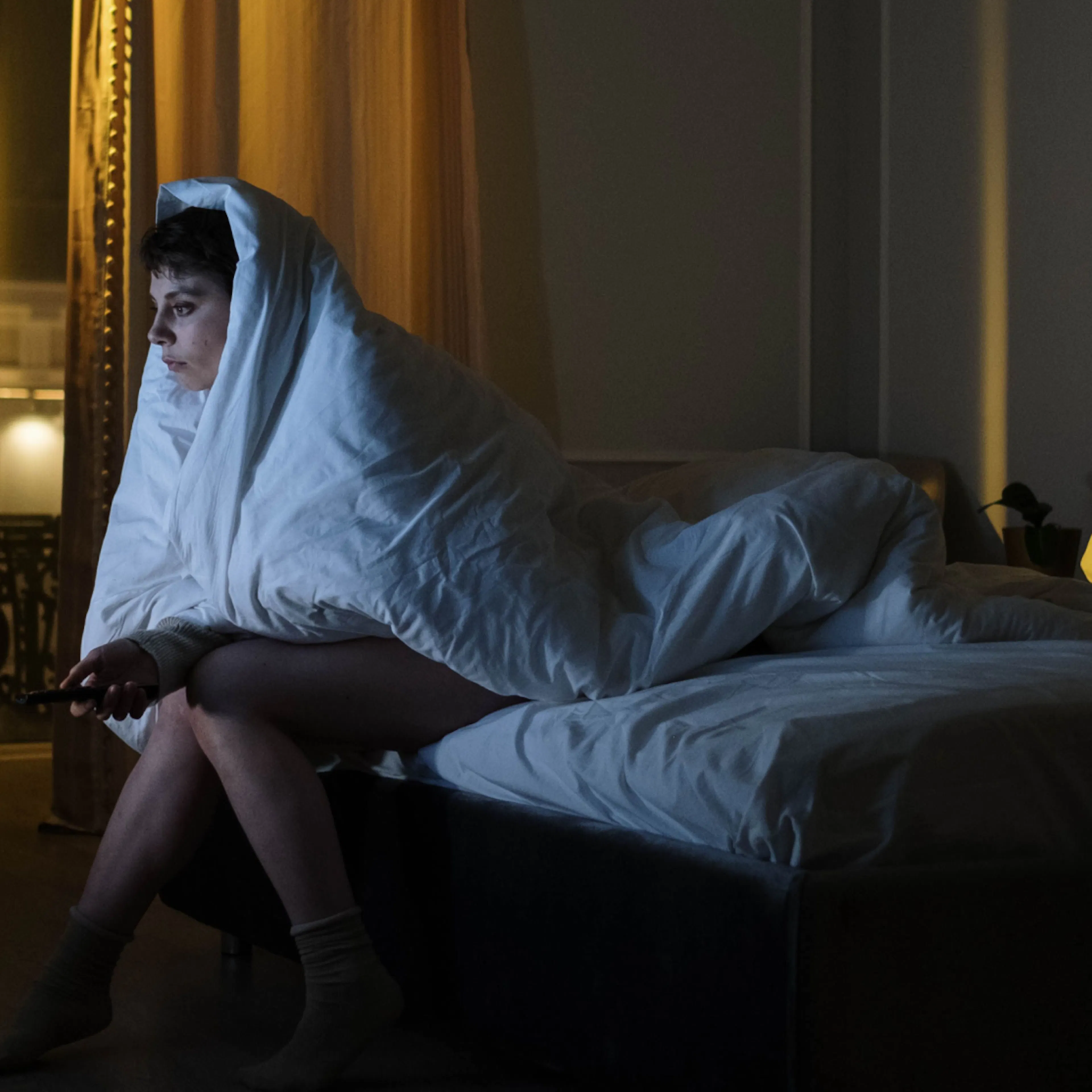 A woman waking up at 3am. She's sitting on the side of her bed with the covers over her head.