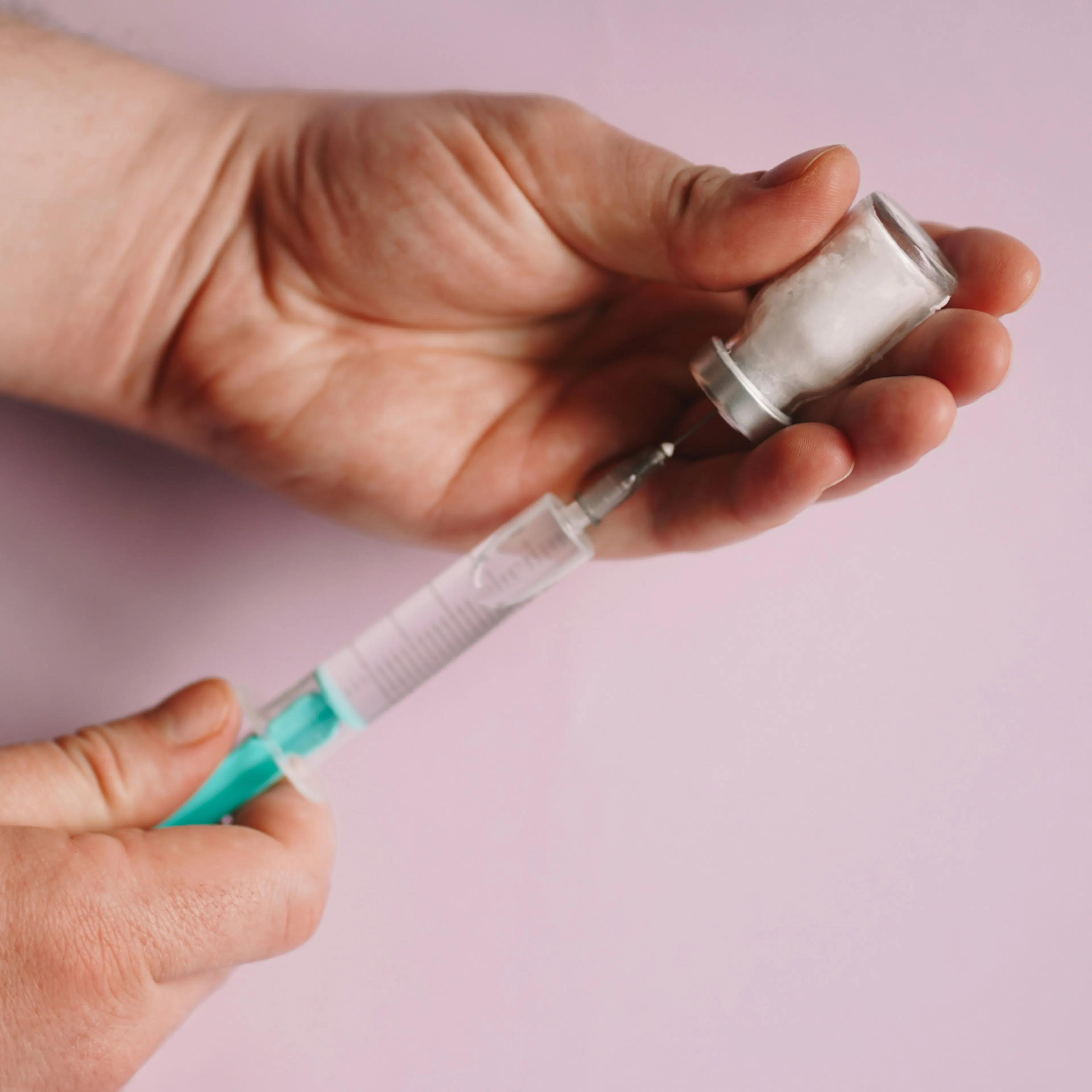 A close-up of a person's hands preparing a medical dose of a weight loss injection by inserting a syringe into a glass vial against a light pink background.