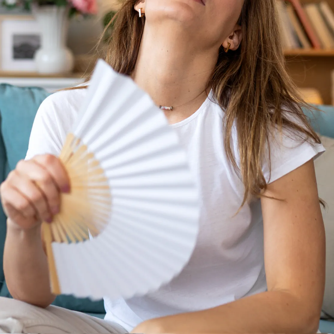 A woman experiencing a hot flash after menopause. She is fanning herself with a fan and looking up.