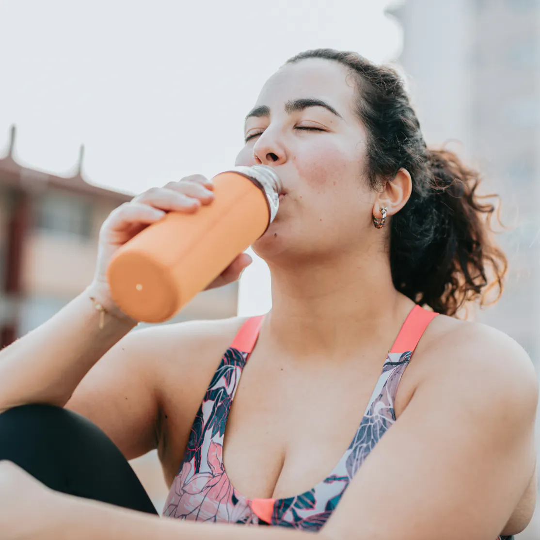 A woman sipping a protein shake from an orange shaker bottle. The bottle is mixed with a protein supplement for women.