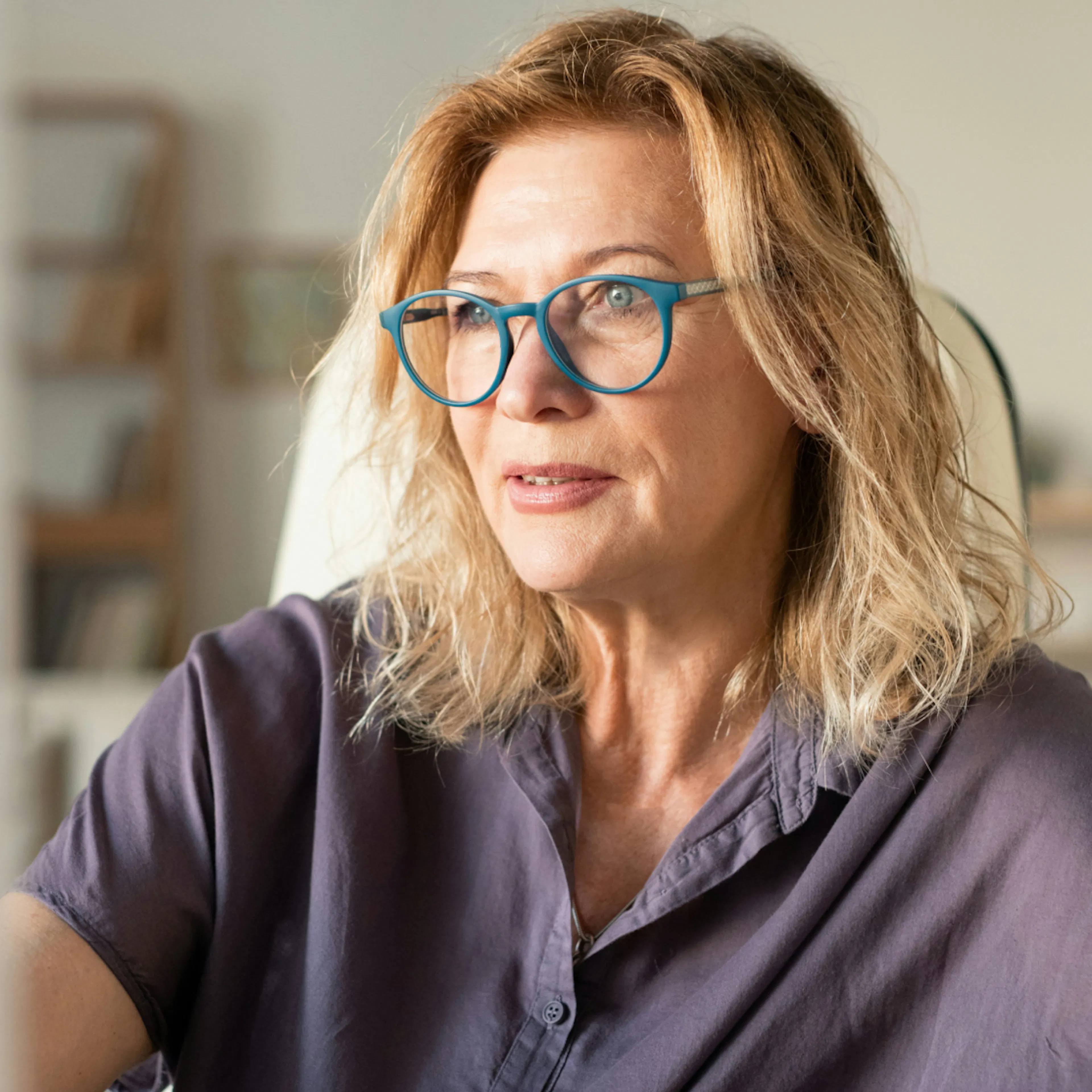 A woman over 50 looking to the side. She's wearing blue glasses and a plum-colored shirt.