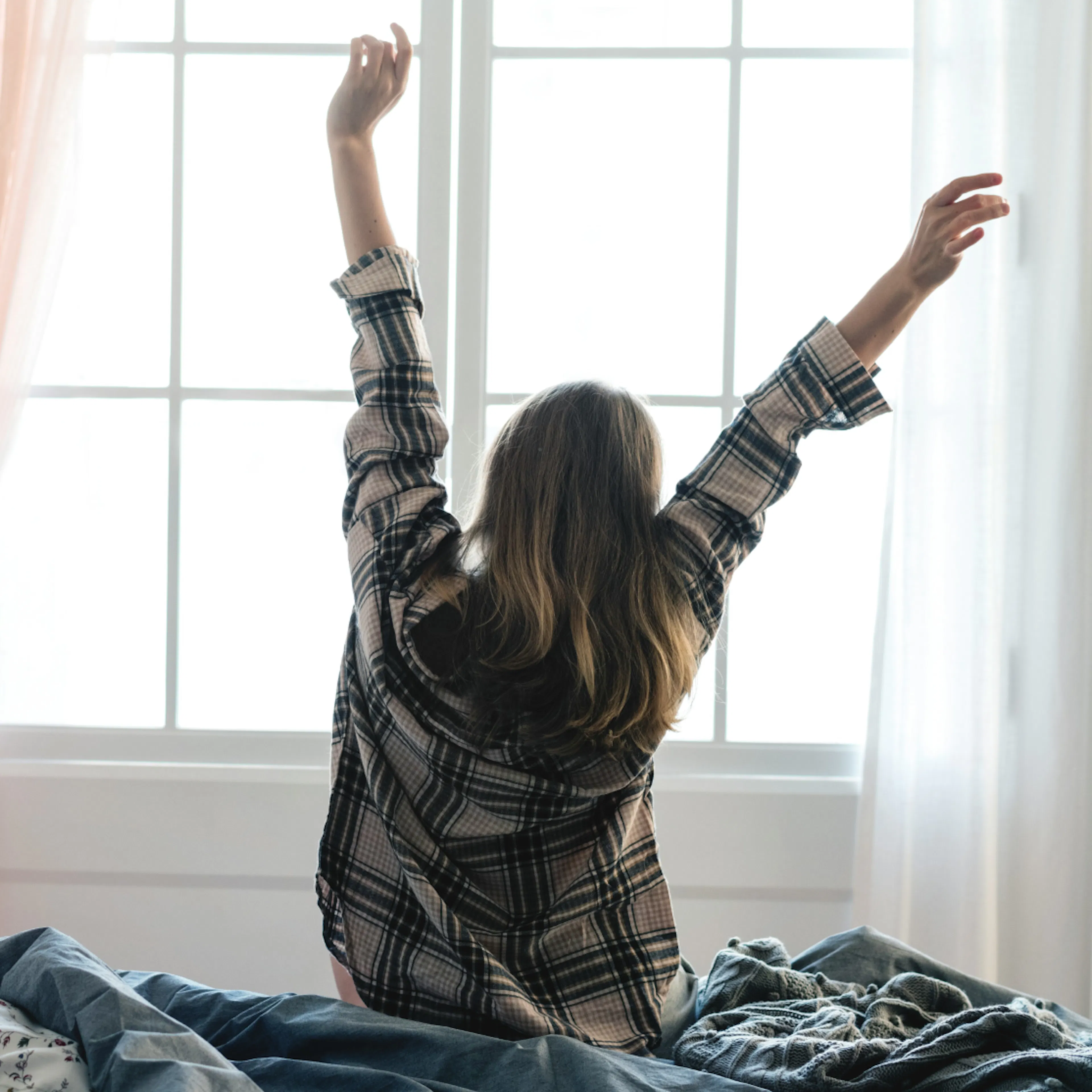 Woman in morning, stretching while sitting in bed, looking out window