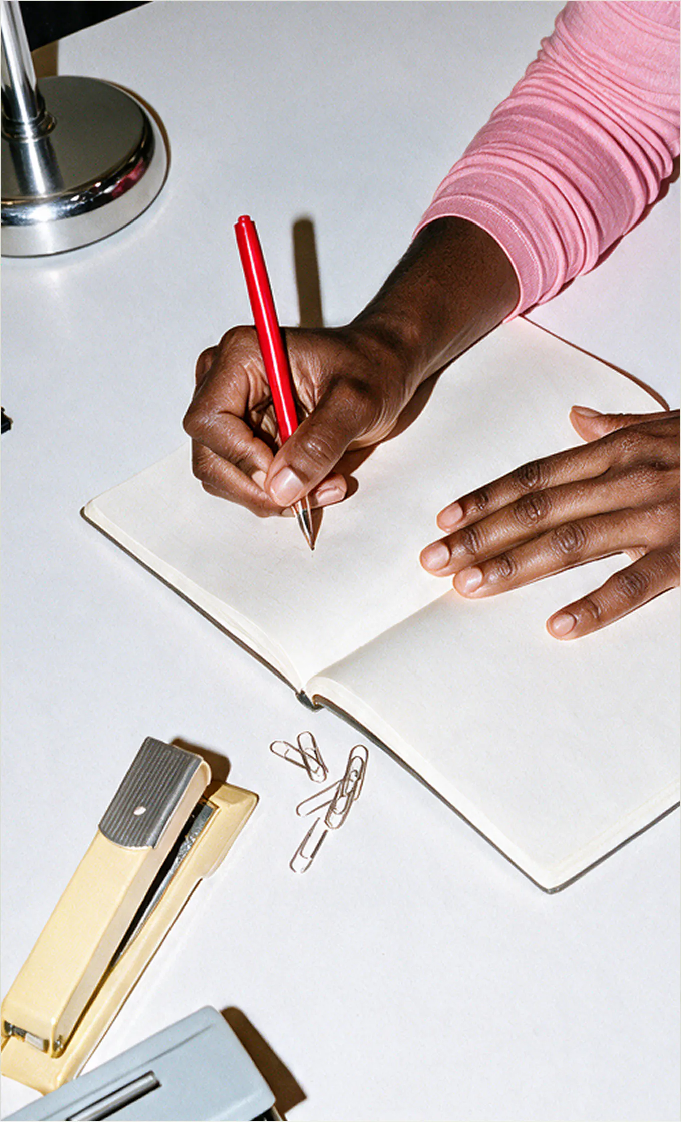 A close-up shot of a woman holding a red pen and writing in an open notebook next to paperclips and a yellow stapler