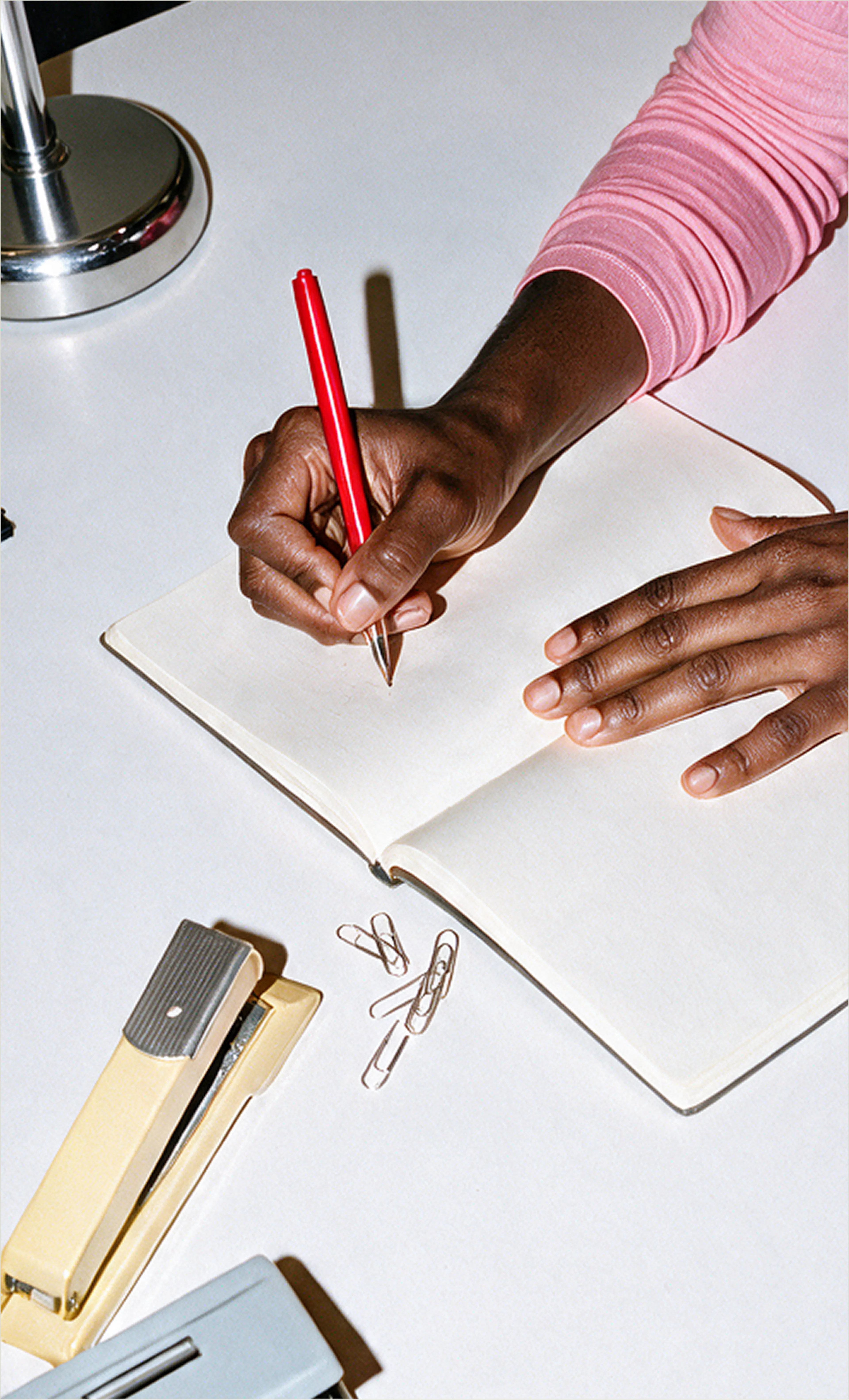 A close-up shot of a woman holding a red pen and writing in an open notebook next to paperclips and a yellow stapler