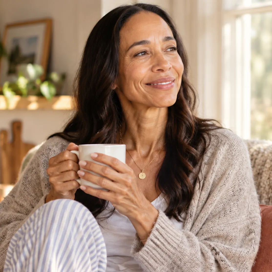 A woman in menopause, sipping her coffee and relaxing at home, smiling and looking into the distance.