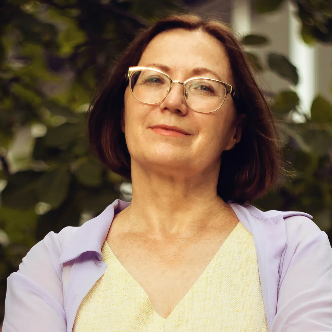 A woman in midlife softly smiling and looking into the camera as she stands outside.