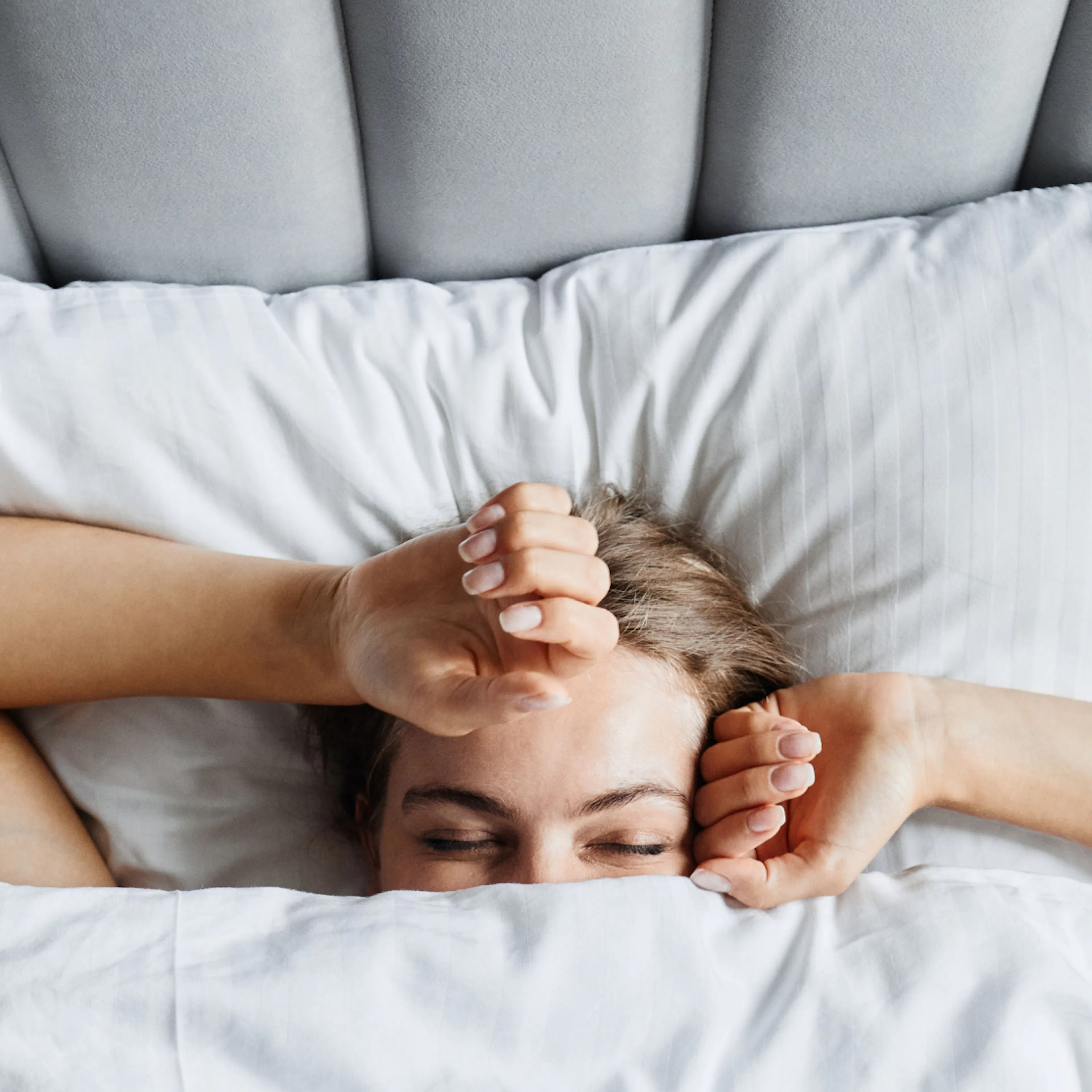 A close-up image of a woman waking up in the morning happy. The covers are up to her eyes and her arms are above her head.