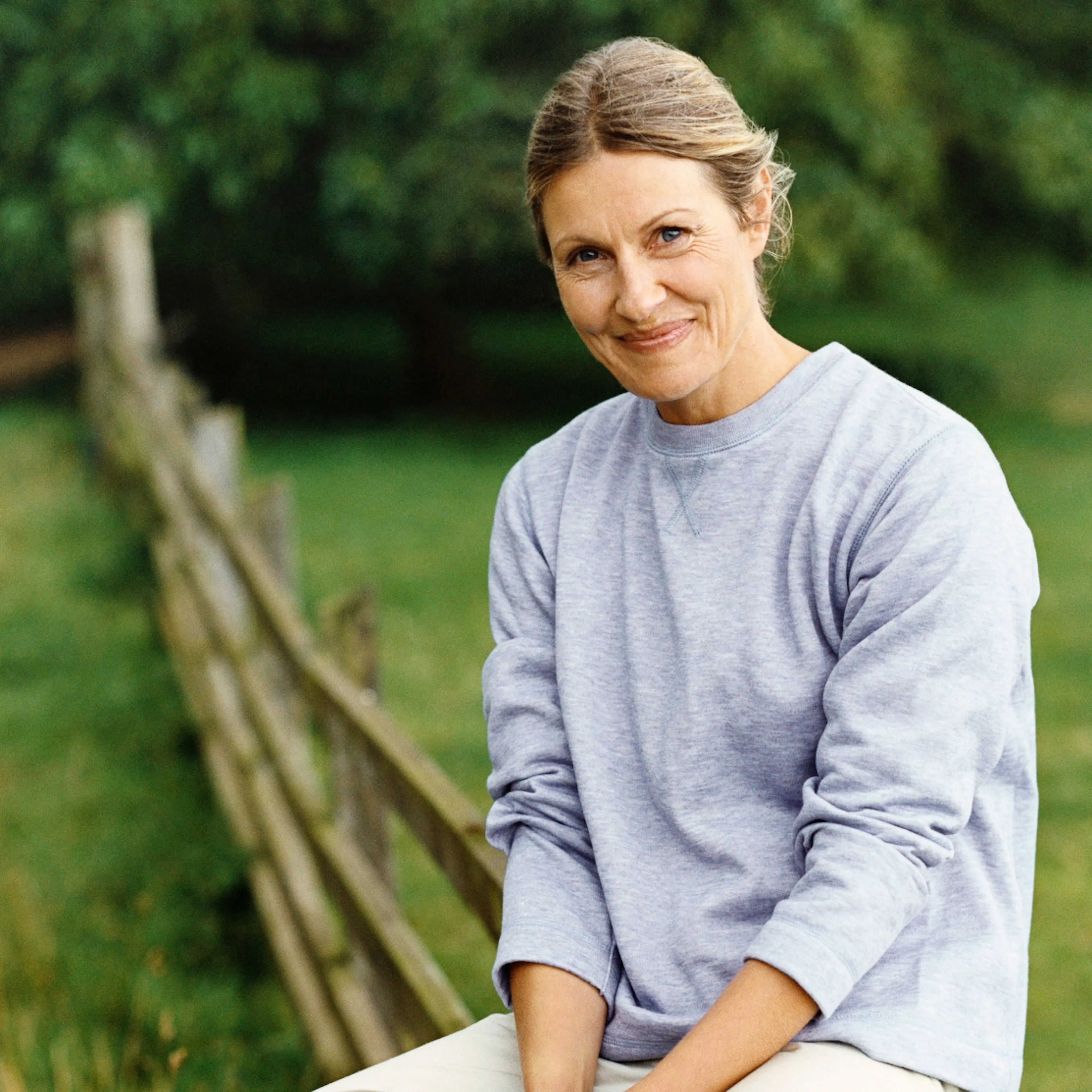 A woman in midlife leaning against a wooden fence outside.
