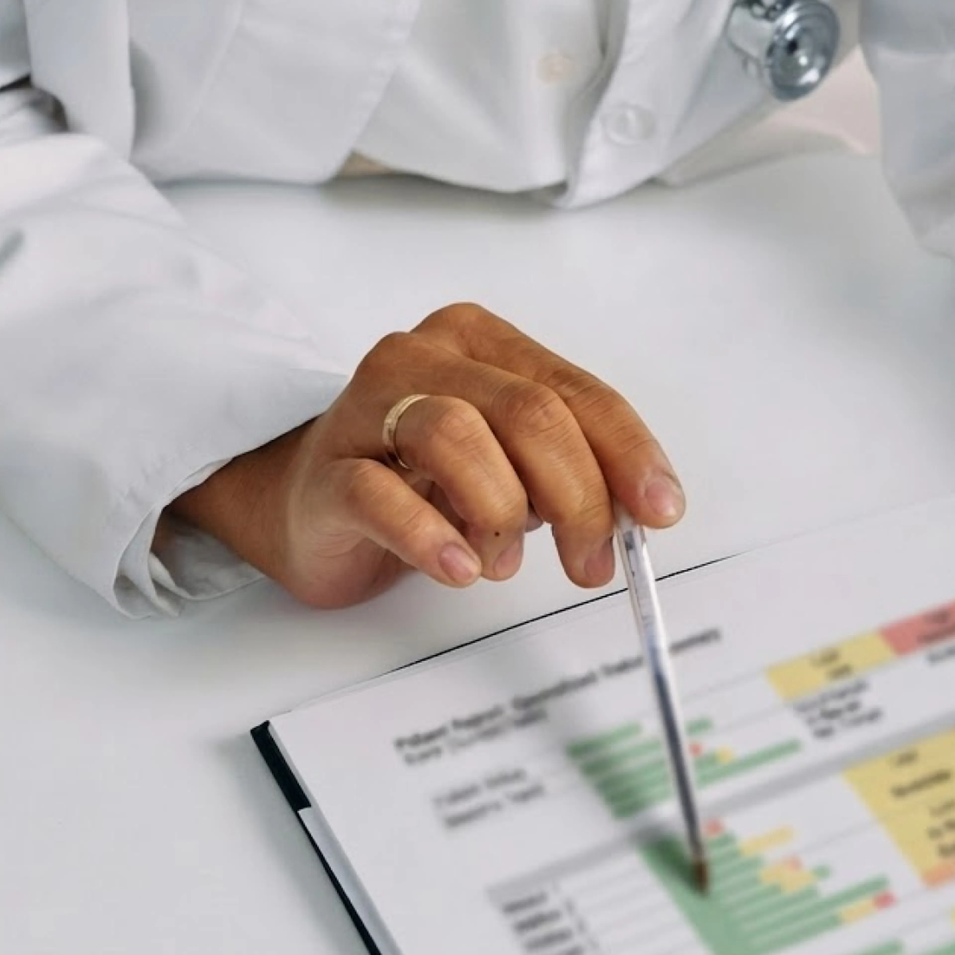 A close-up of a clinician's hand with a pen pointed to a blurred-out medical document.