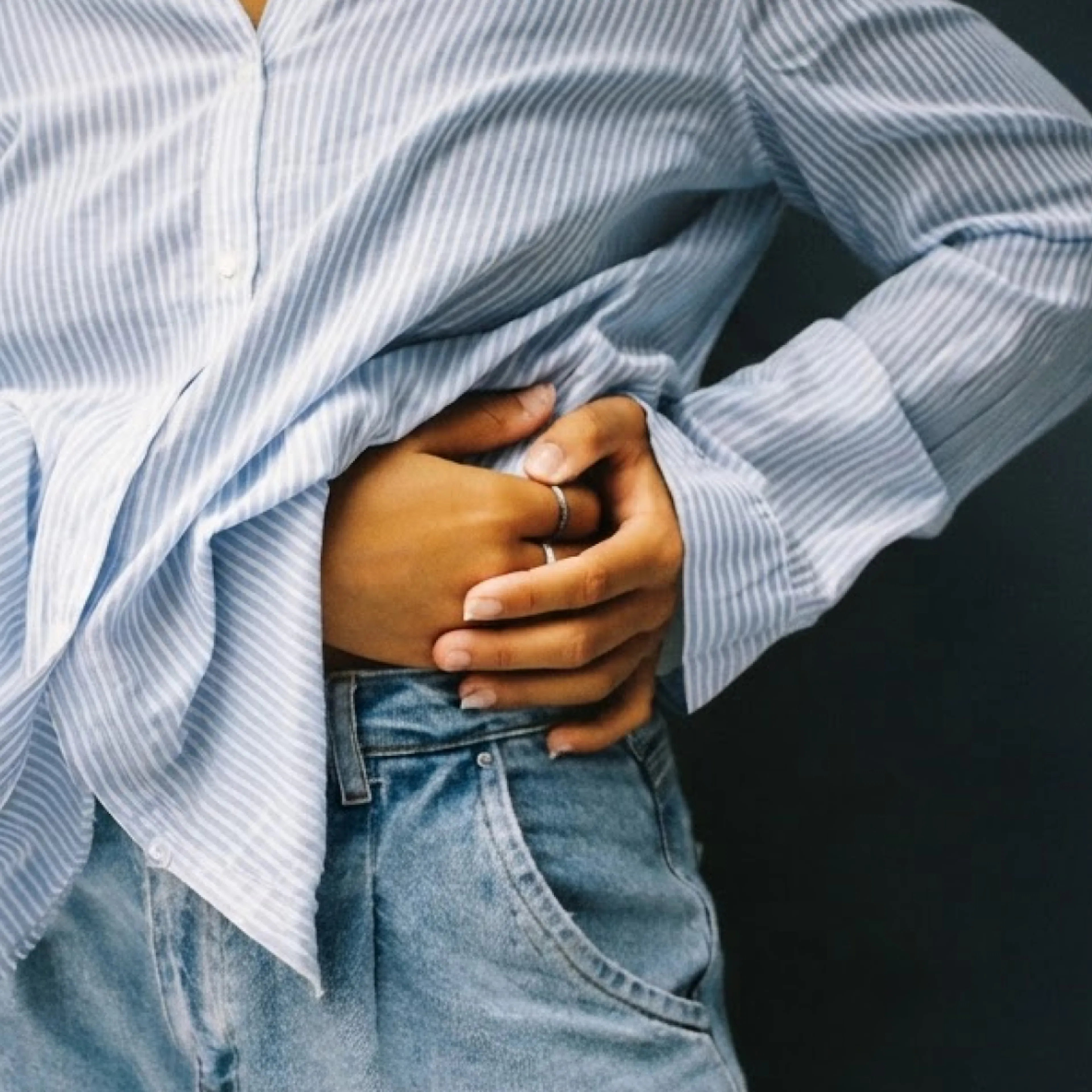 A close-up image of a woman in midlife holding her belly. She's wearing jeans and a blue-striped button down.