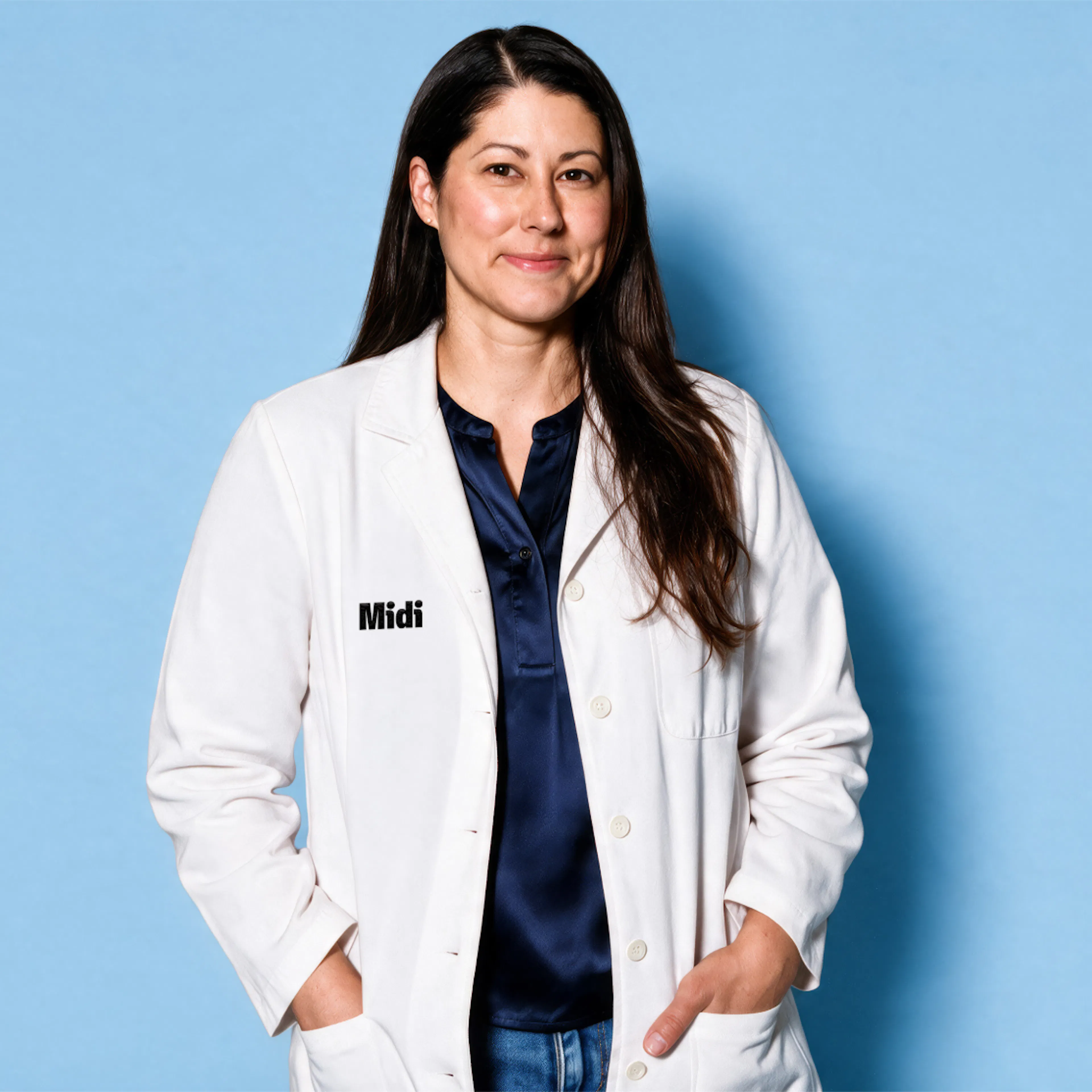 A photo of a Midi clinician standing and smiling, wearing a white lab coat and a navy top.