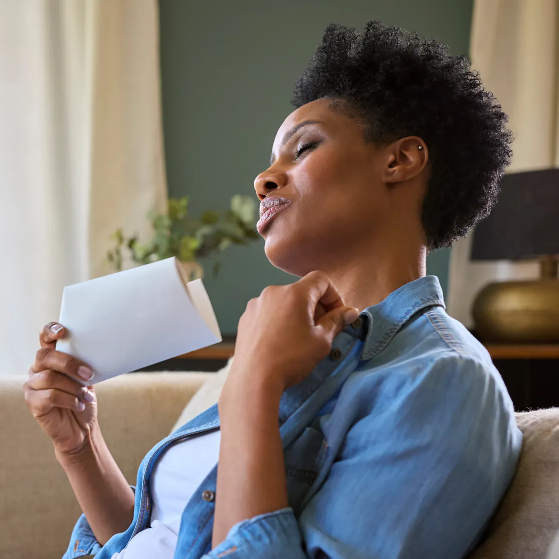 A woman in midlife experiencing a hallmark symptom of menopause, hot flashes. She is fanning herself off with a piece of paper while sitting on the couch inside.