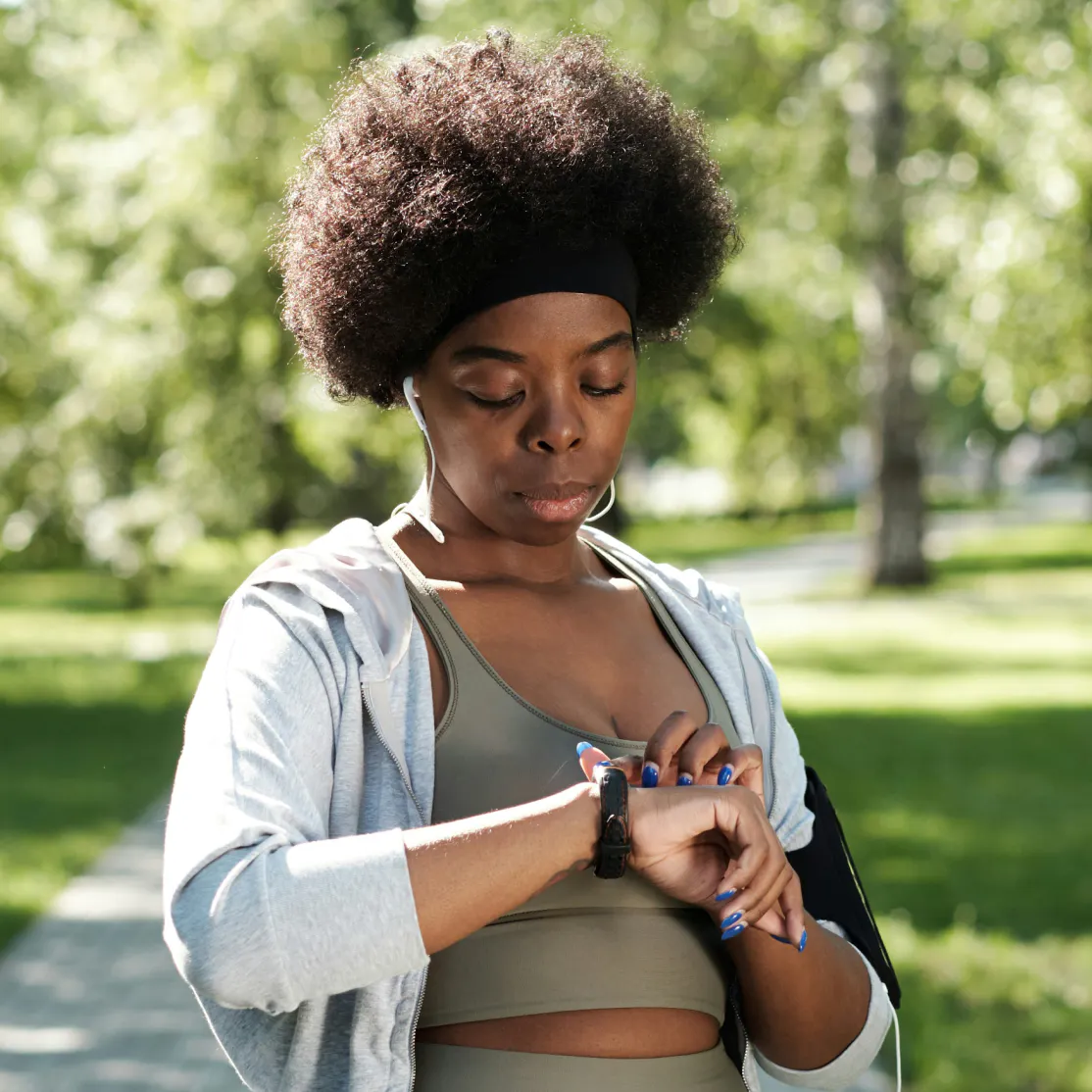 A woman in midlife in workout clothes, standing outside, looking at her watch.