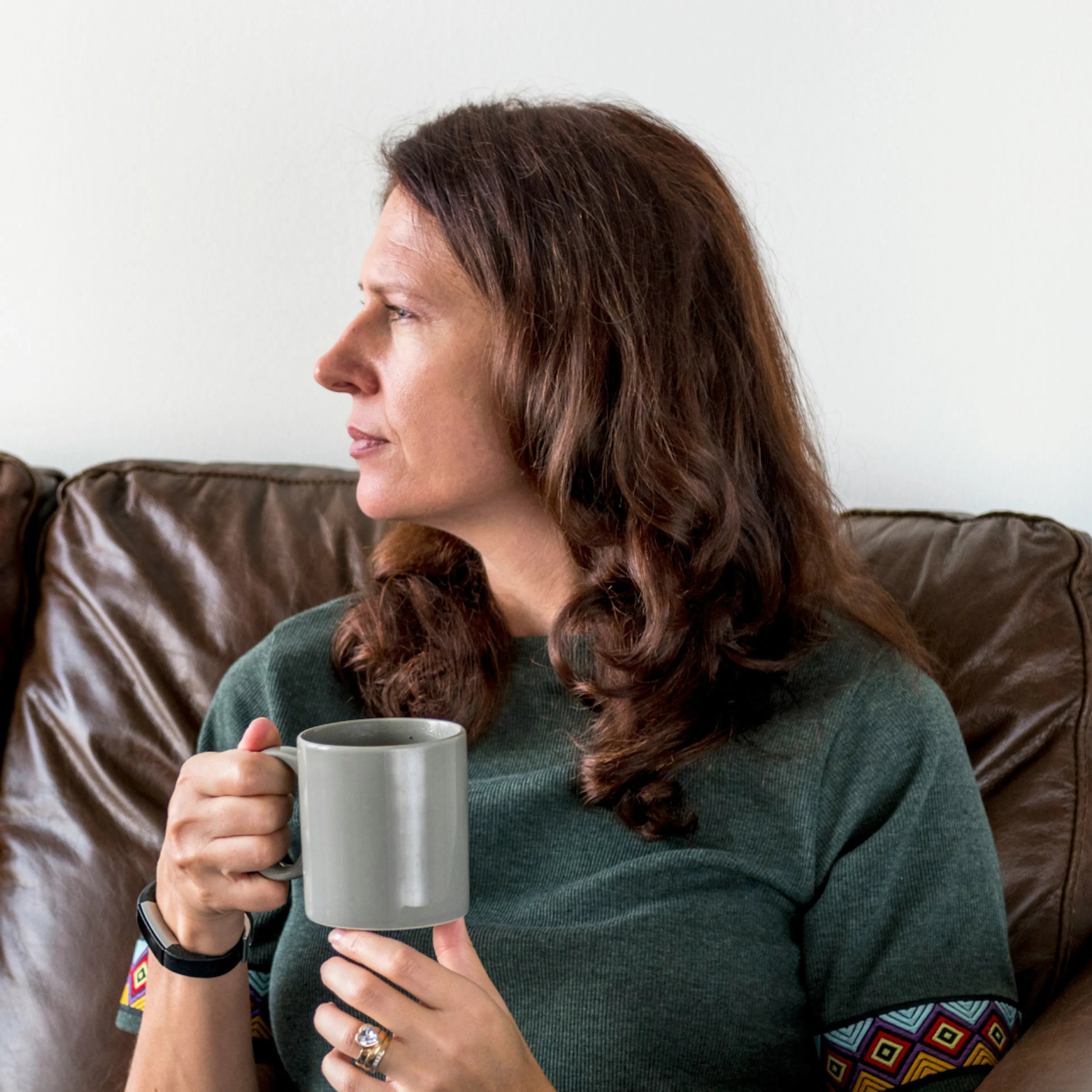 A woman in midlife holding a cup of coffee while sitting on the couch and looking to her right.