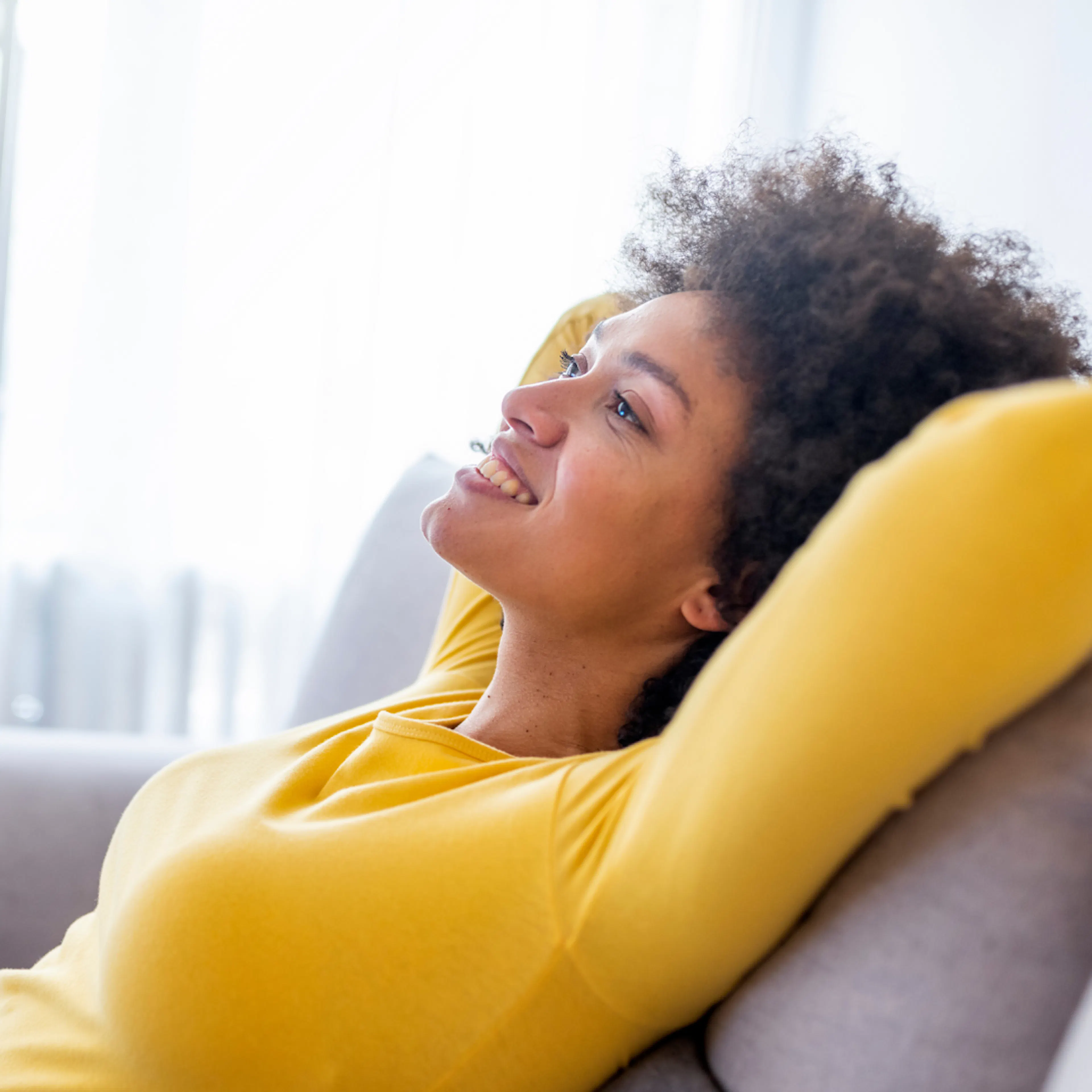 A woman in a yellow shirt, kicking back and relaxing at home, smiling and not feeling stressed at all.