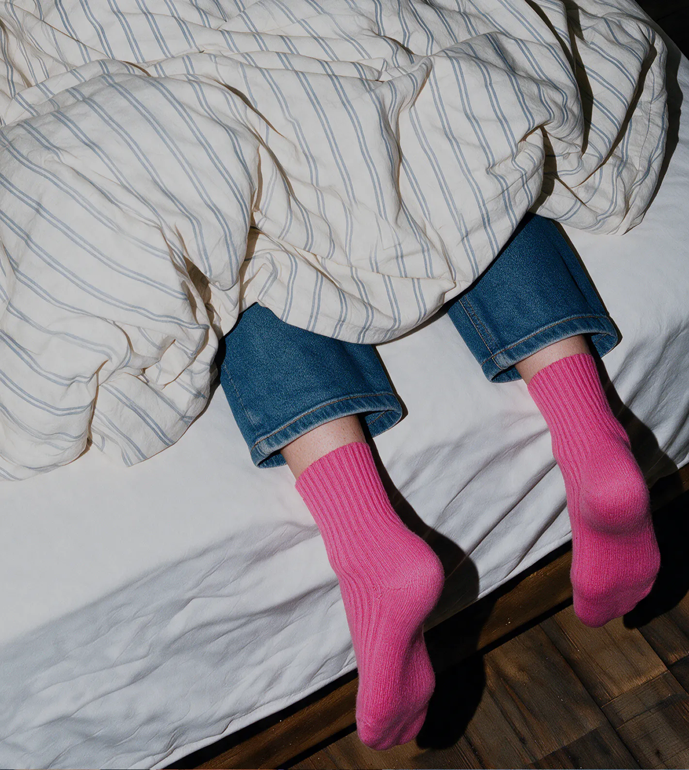 A person wearing blue jeans and bright pink ribbed knit socks resting their legs on a bed