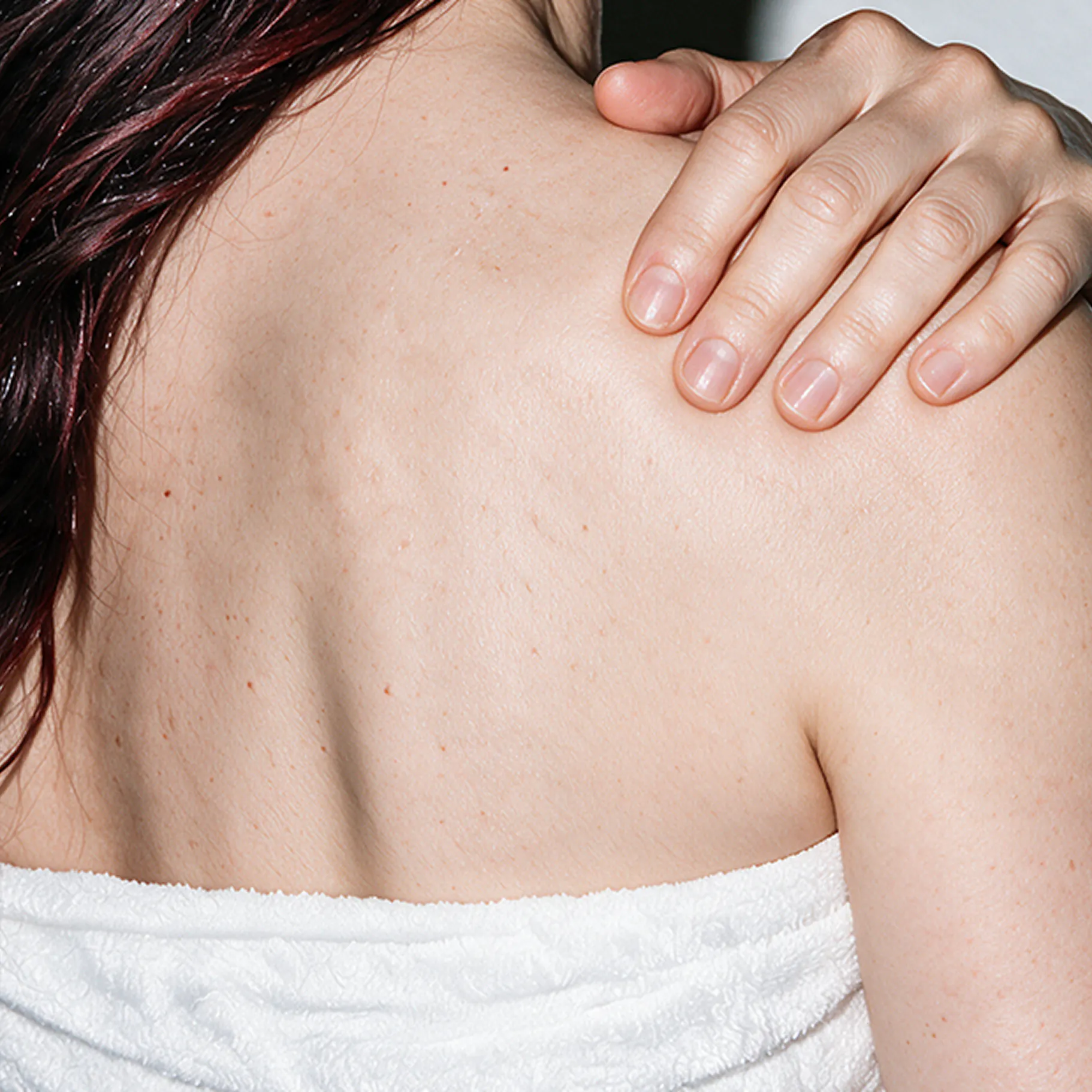 A rear view of a person wearing a white towel, holding their own shoulder with one hand