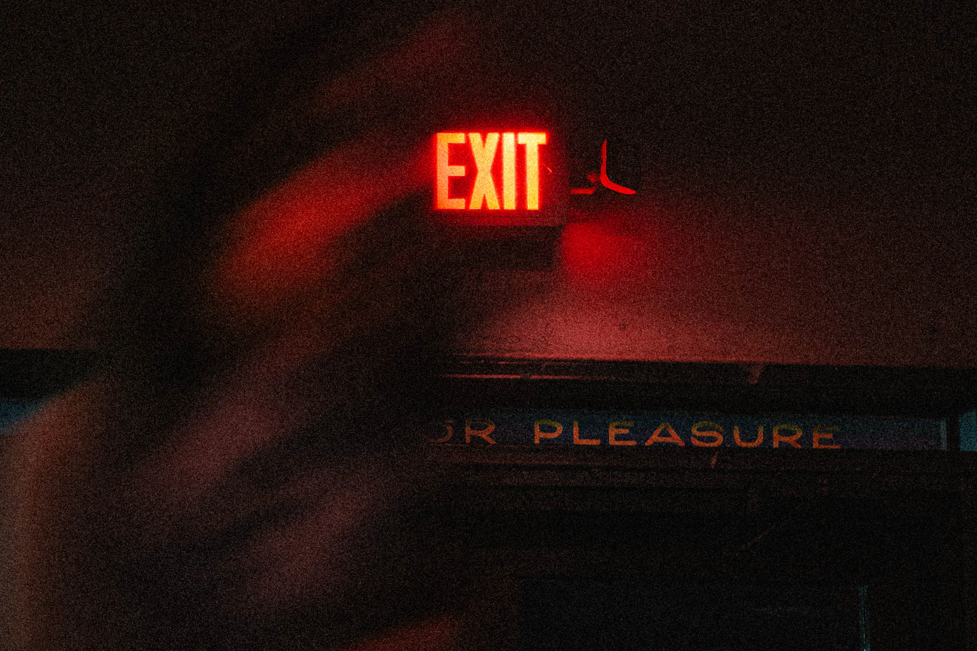 A neon exit sign above a door that says pleasure with a woman through it. Exit for Pleasure from Jonathan Grado's Dirty Static.