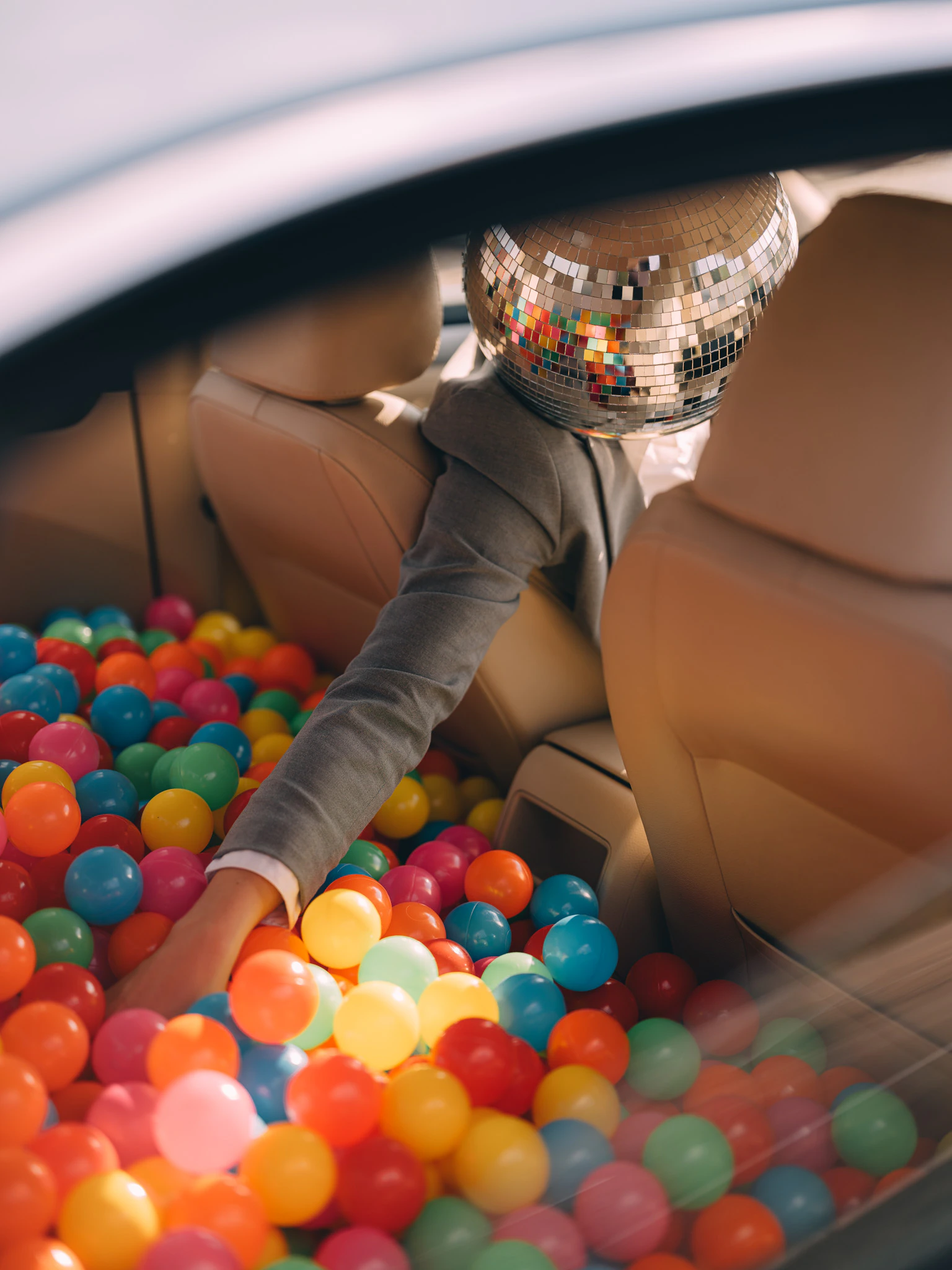 Looking through a car window at a person in a suit and disco ball for a head reaching into a ball pit in the back seat. For Internal Use Only from Jonathan Grado's Happy Hour series.