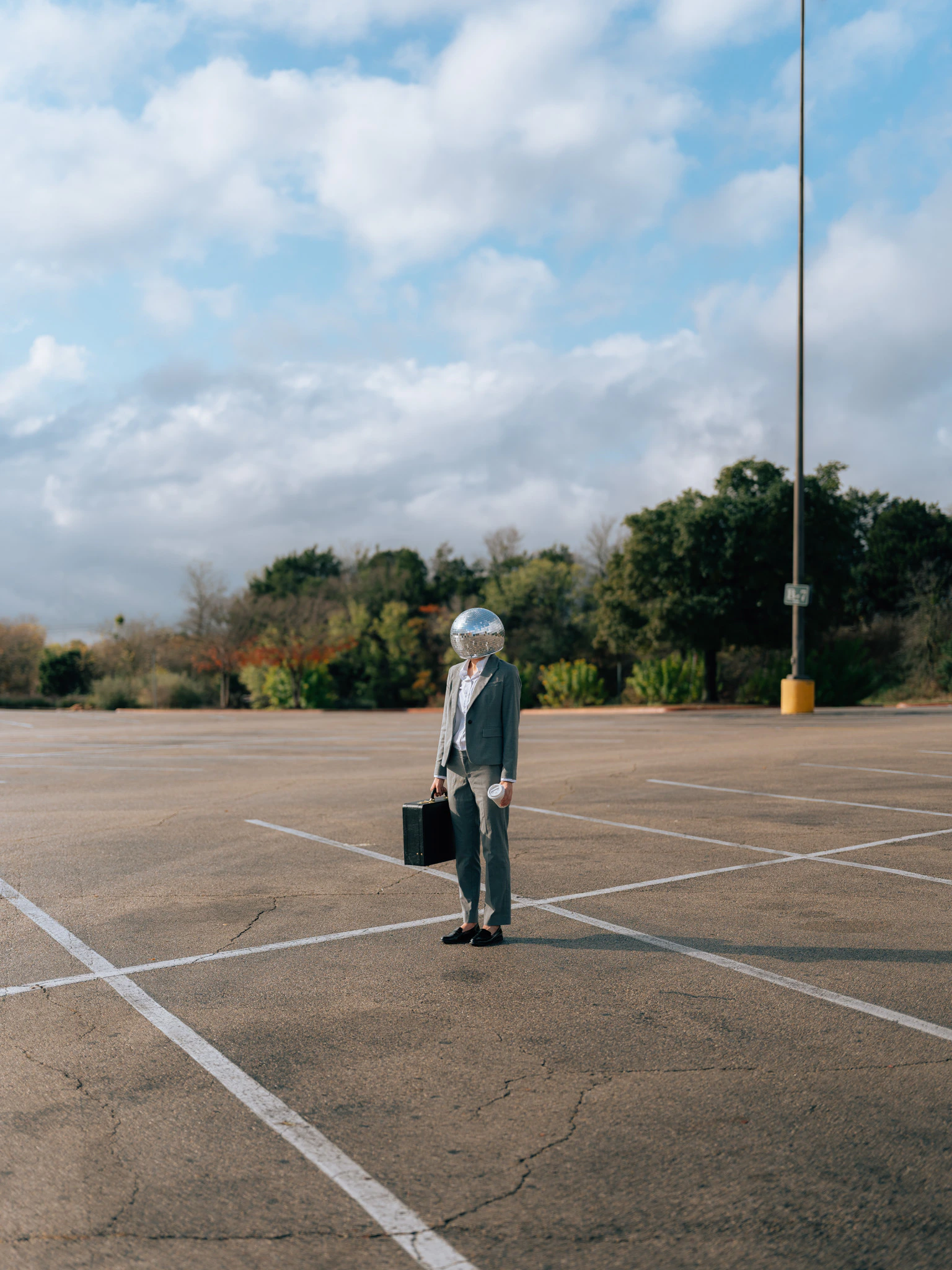 A disco ball headed person with a briefcase in an empty parking lot. Performance Review from Jonathan Grado's Happy Hour series.