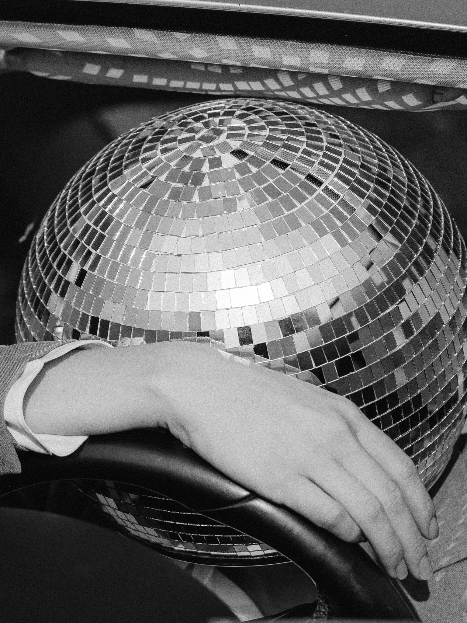 A disco ball head resting on a car steering wheel and hand in black and white. Q4 from Jonathan Grado's Happy Hour series.