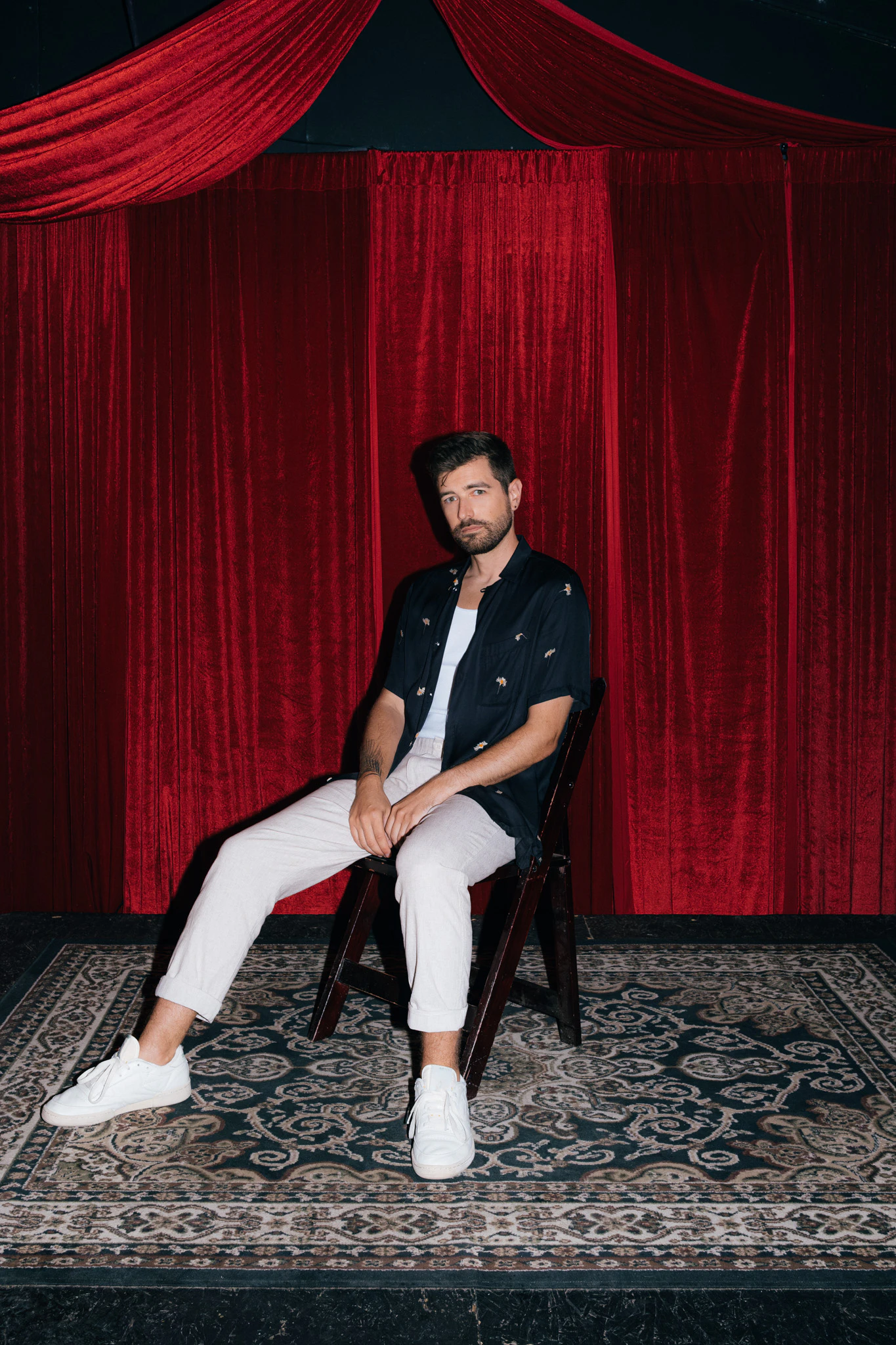 Jonathan Grado sitting on a chair on a red velvet stage wearing an open dandelion shirt and white sneakers.
