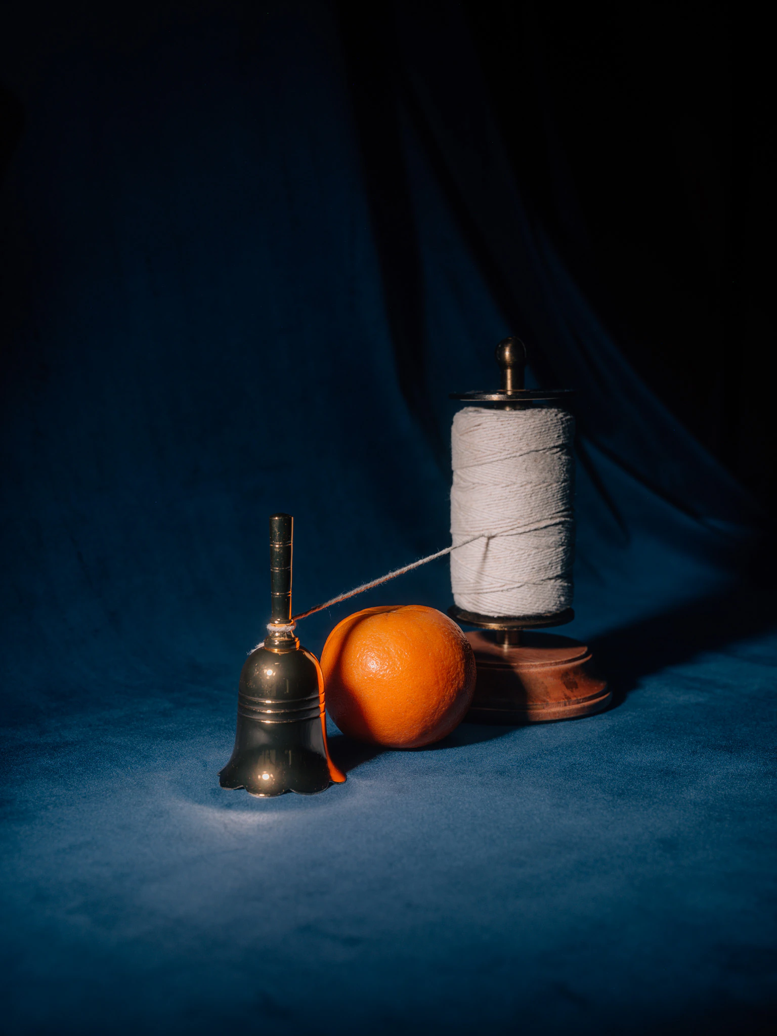 An orange under a thread connecting the spool to a hand bell. Risk Aversion from Jonathan Grado's Saint Citrus series.
