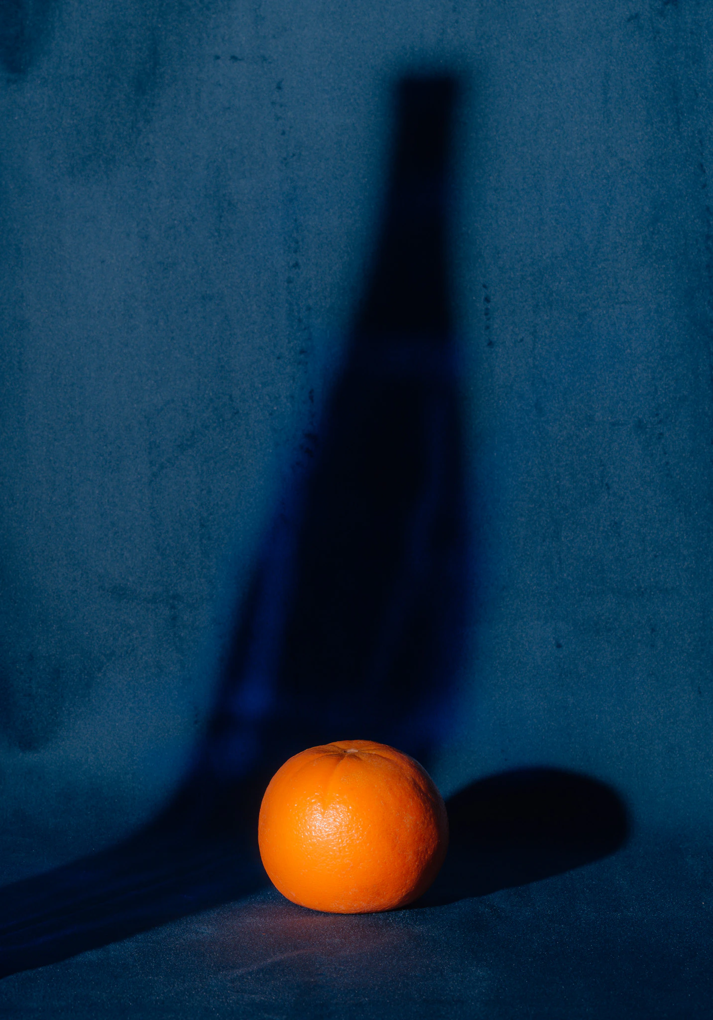 An orange with the shadow of a glass bottle on blue velvet. Imposition from Jonathan Grado's Saint Citrus series.
