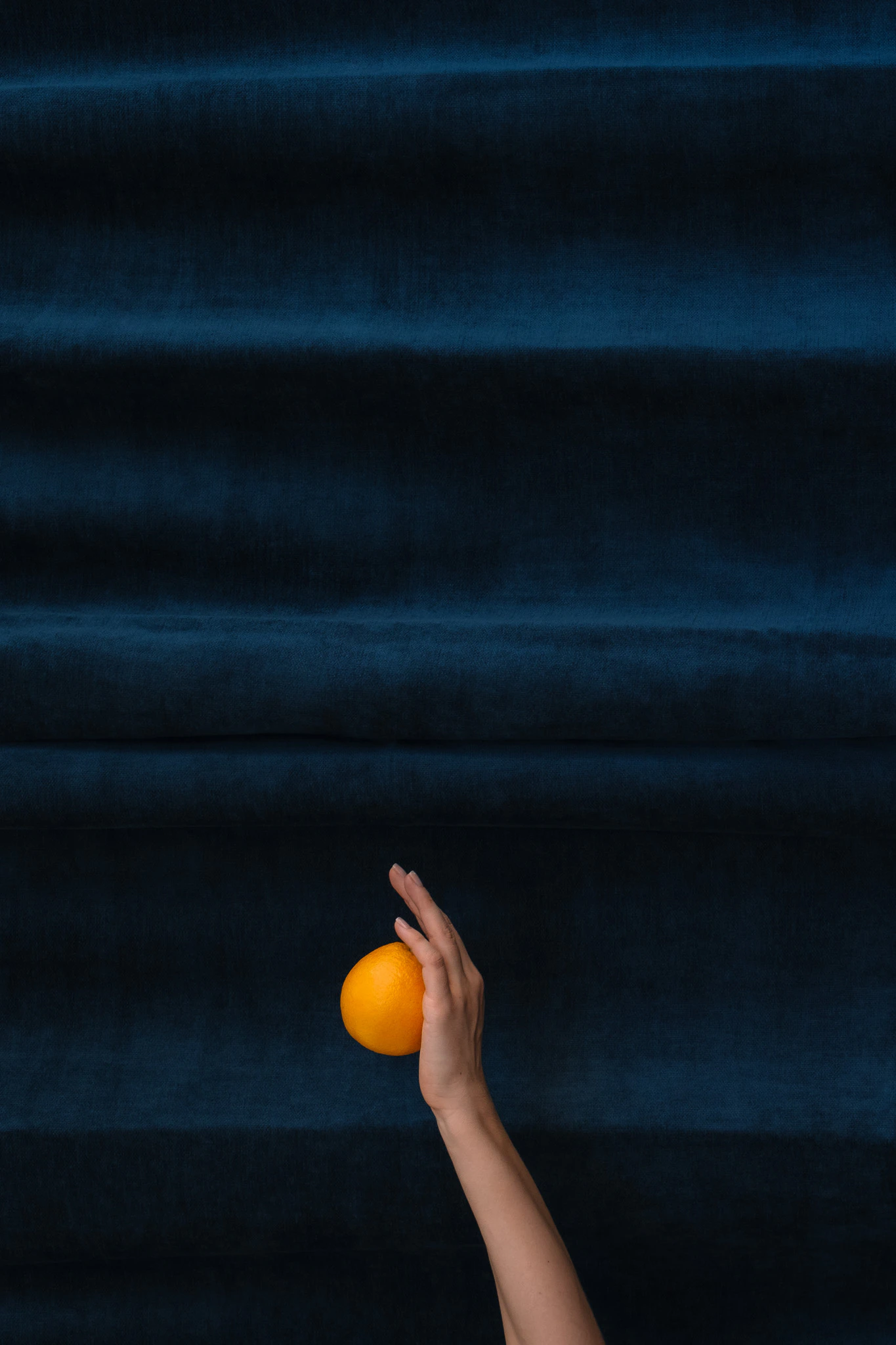 The hand of Saint Citrus holding an orange upwards in front of blue velvet. Saint Citrus #01 from Jonathan Grado's Saint Citrus series.
