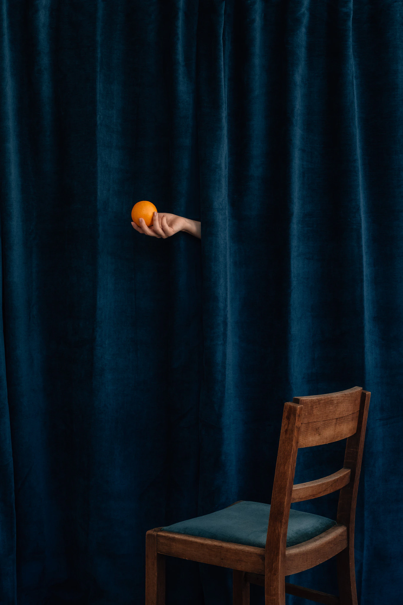 The hand of Saint Citrus holding an orange in front of blue velvet above an empty chair. Saint Citrus #02 from Jonathan Grado's Saint Citrus series.