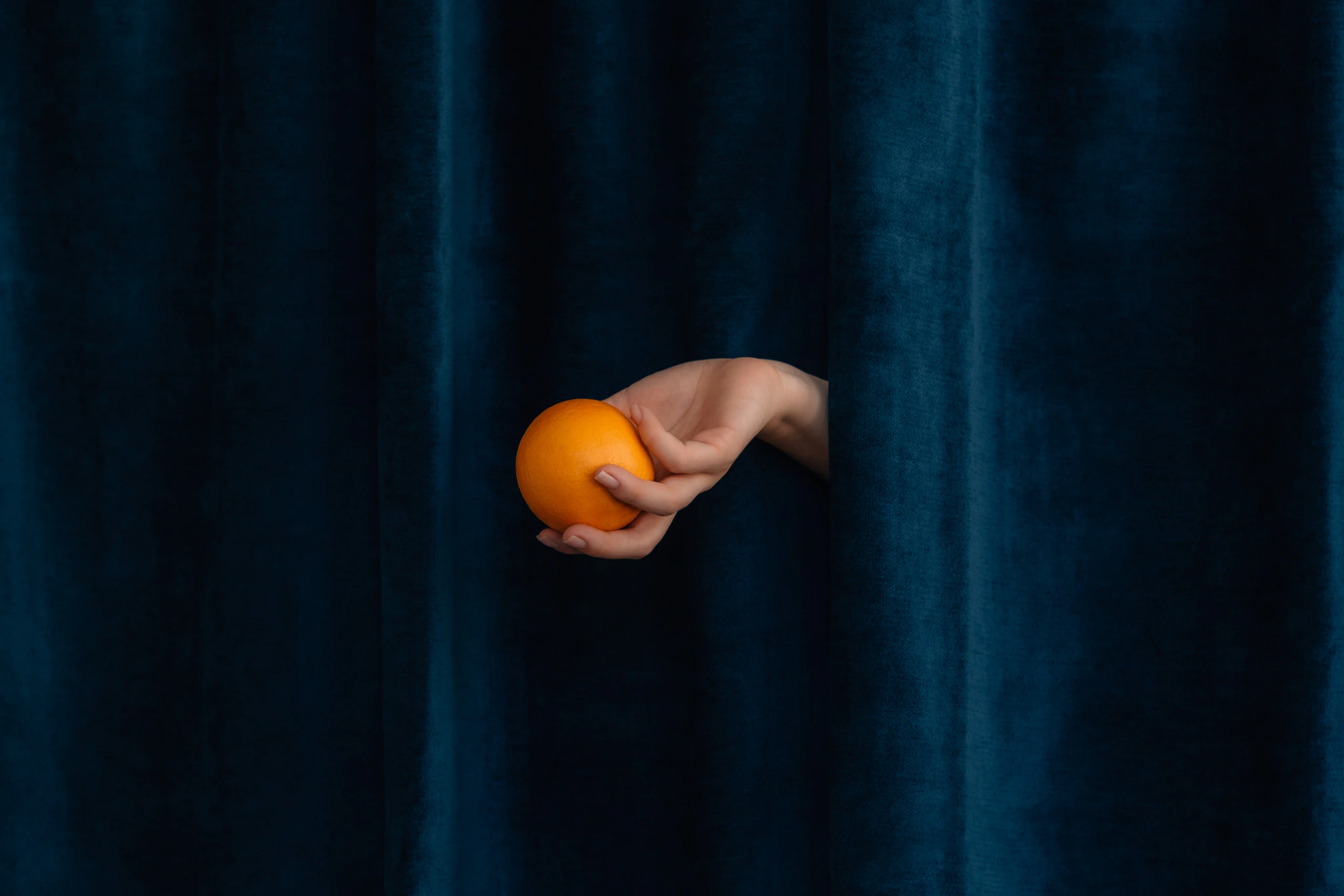 The hand of Saint Citrus holding an orange in front of blue velvet. Saint Citrus #03 from Jonathan Grado's Saint Citrus series.