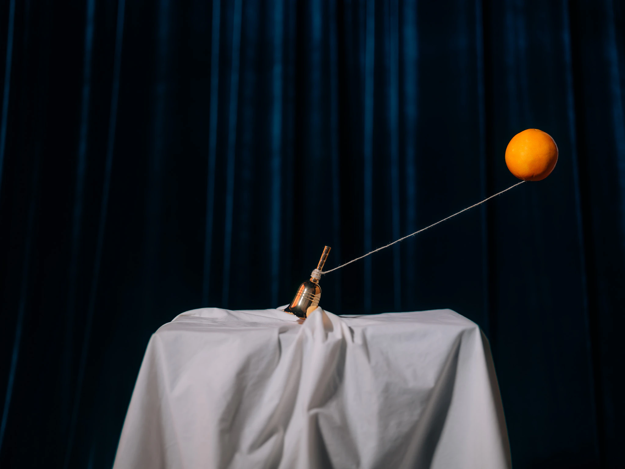A small golden hand bell being pulled by a floating orange on a string. The Idea #01 from Jonathan Grado's Saint Citrus series.