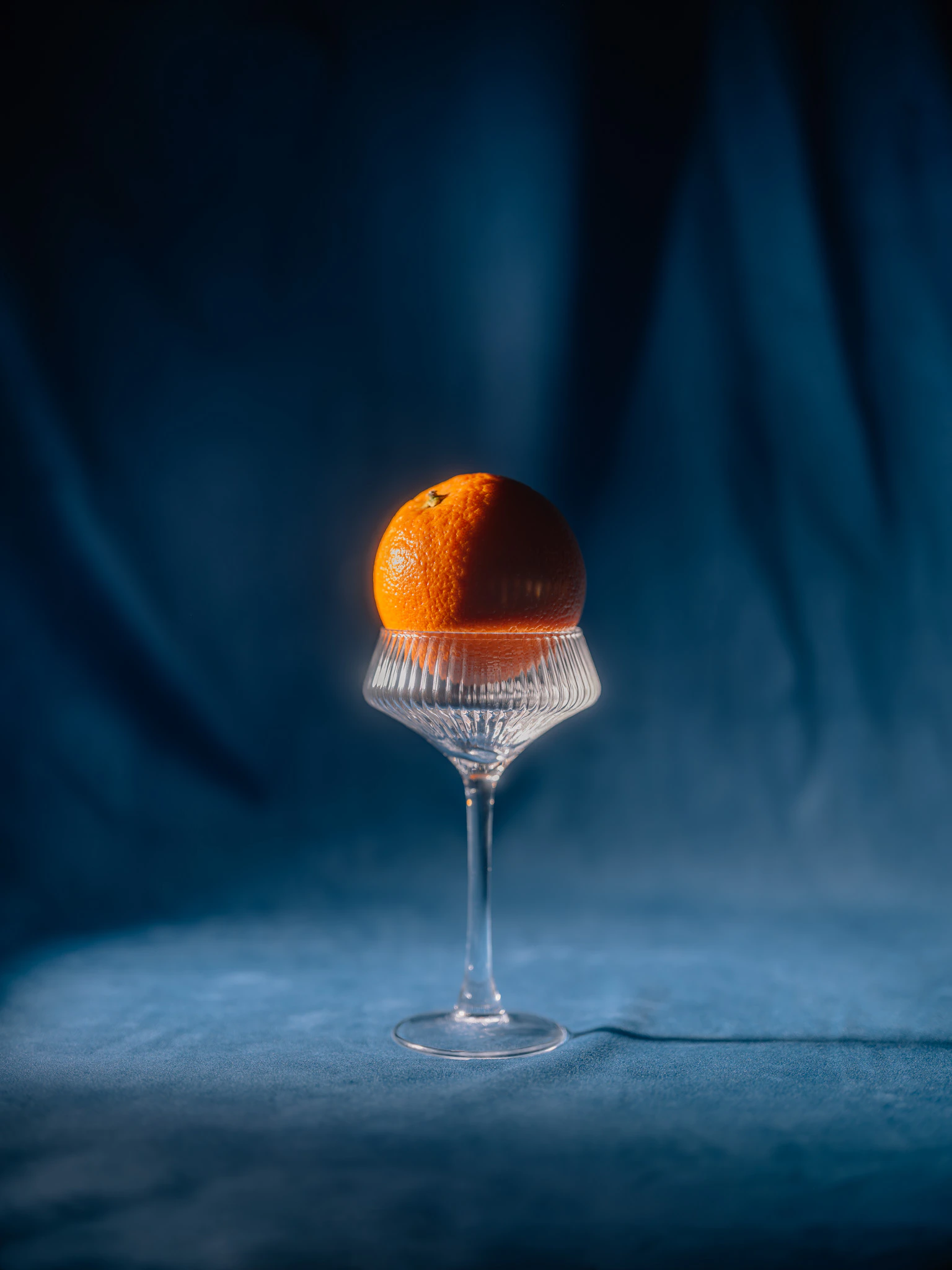 An orange inside a drinking glass on blue velvet. Zero Proof of Concept from Jonathan Grado's Saint Citrus series.