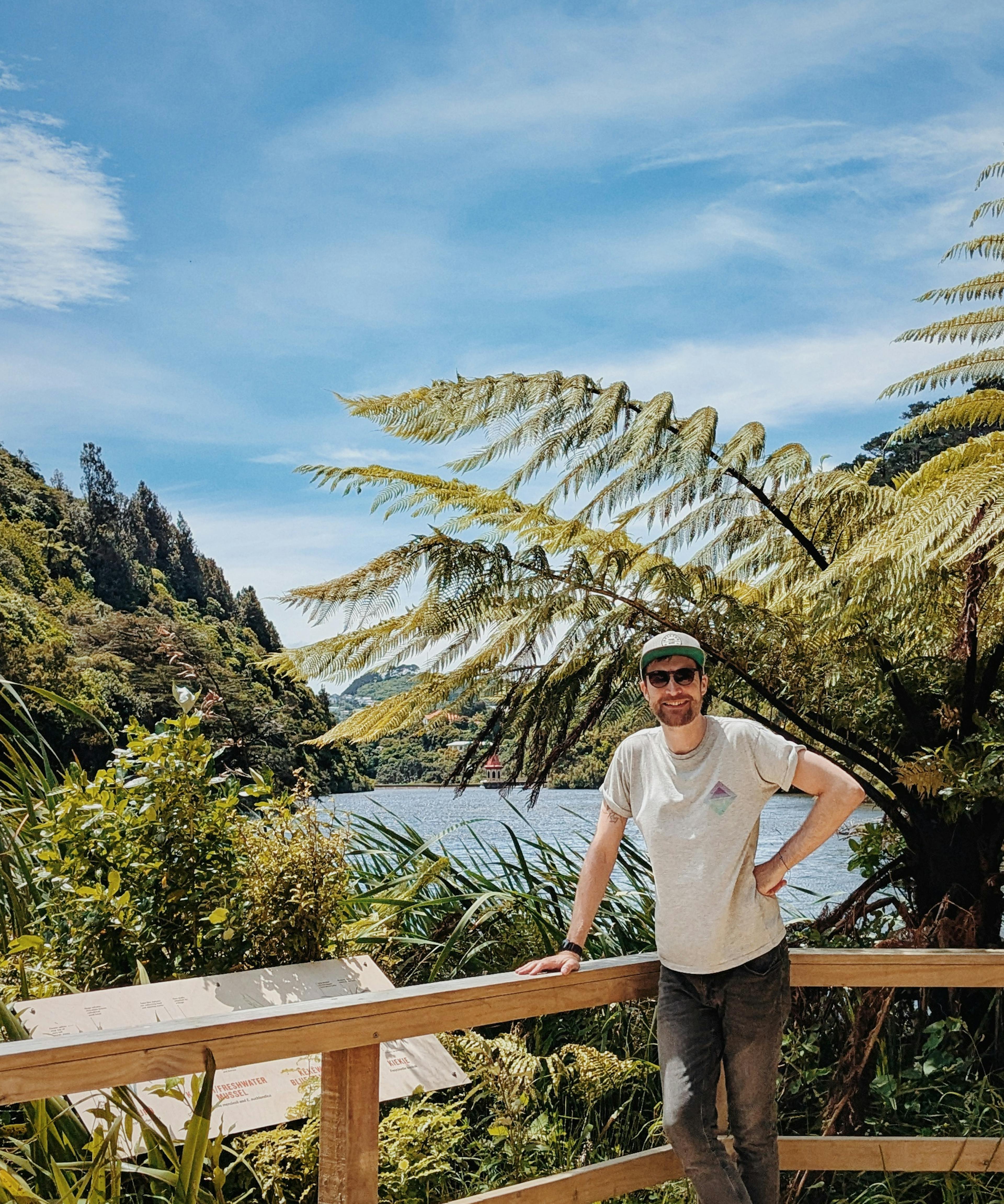 Jon stands in Zealandia, in Wellington, New Zealand. There are wispy clouds in the sky across a blue, sunny sky. The jungle stretches off in the background with a lake in the centre.