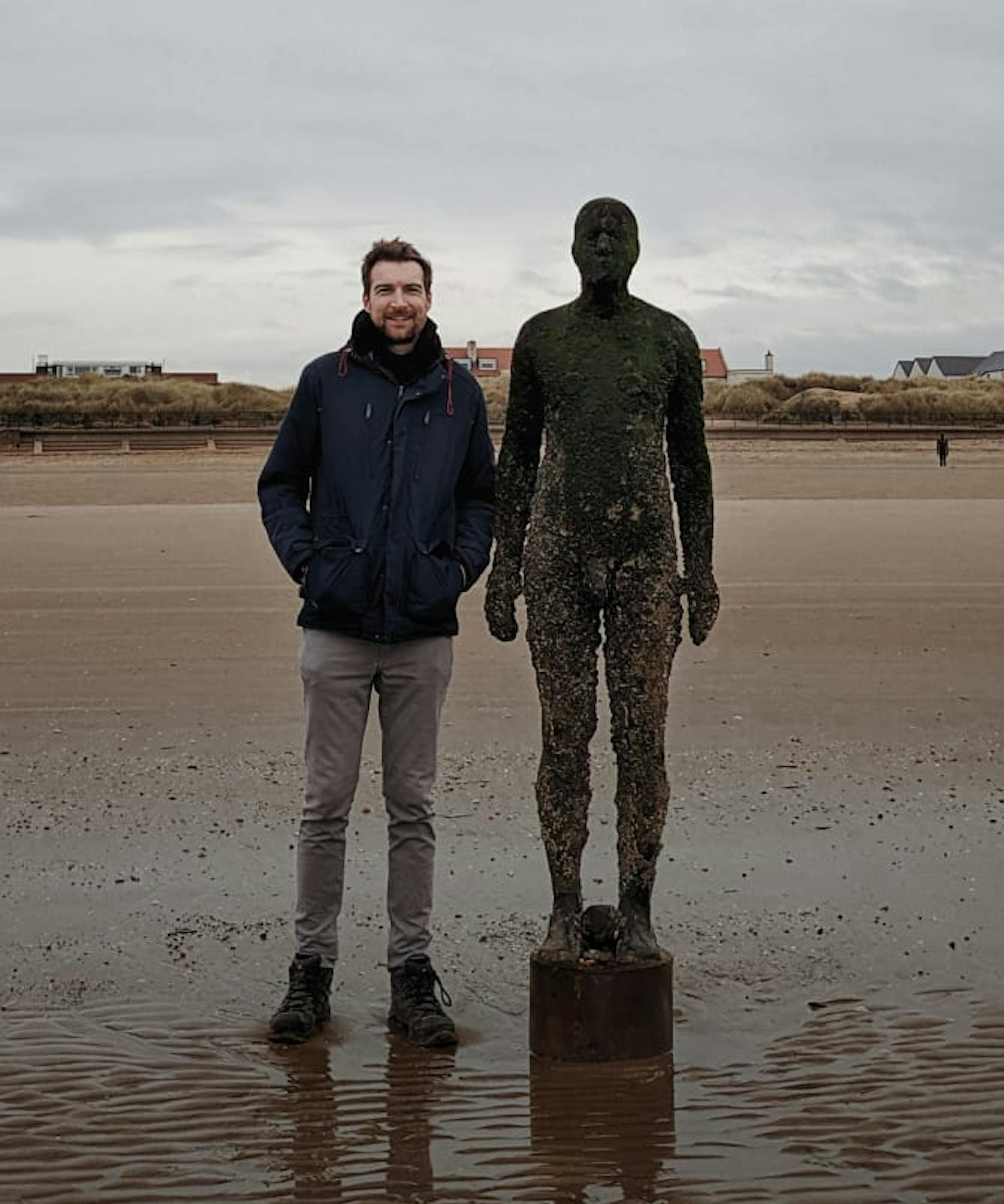 Jon stands on Crosby Beach next to a statue from Antony Gormley's "Another Place"