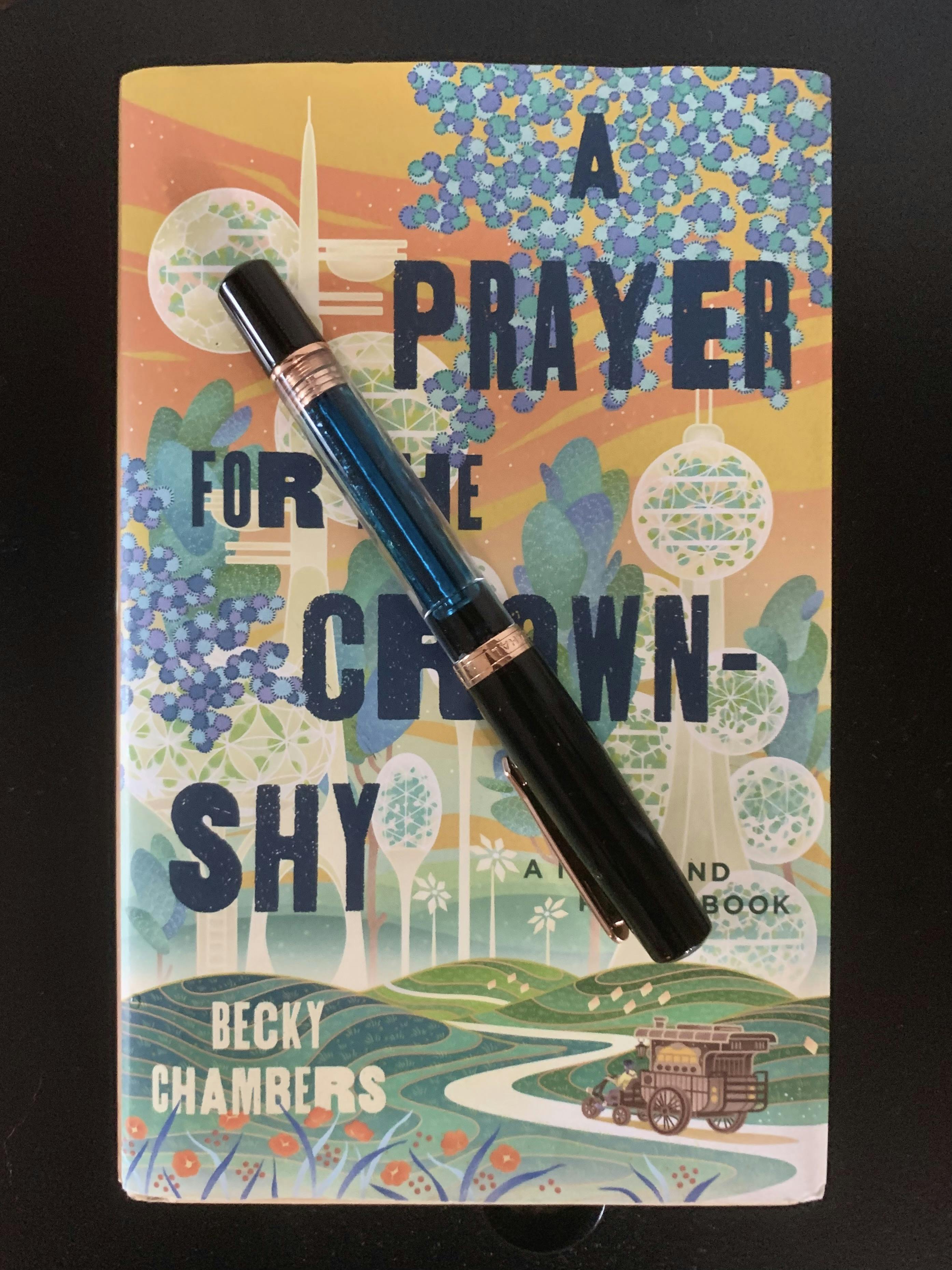 A fountain pen rests on the cover a book. The pen is a Nahvalur Original Plus, a partially-transparent 'demonstrator' pen. The book is 'A Prayer for the Crown-Shy' by Becky Chambers. The gold, blue, and green color scheme of the cover illustration matches the gold banding on the pen and the turquoise ink it's filled with. 