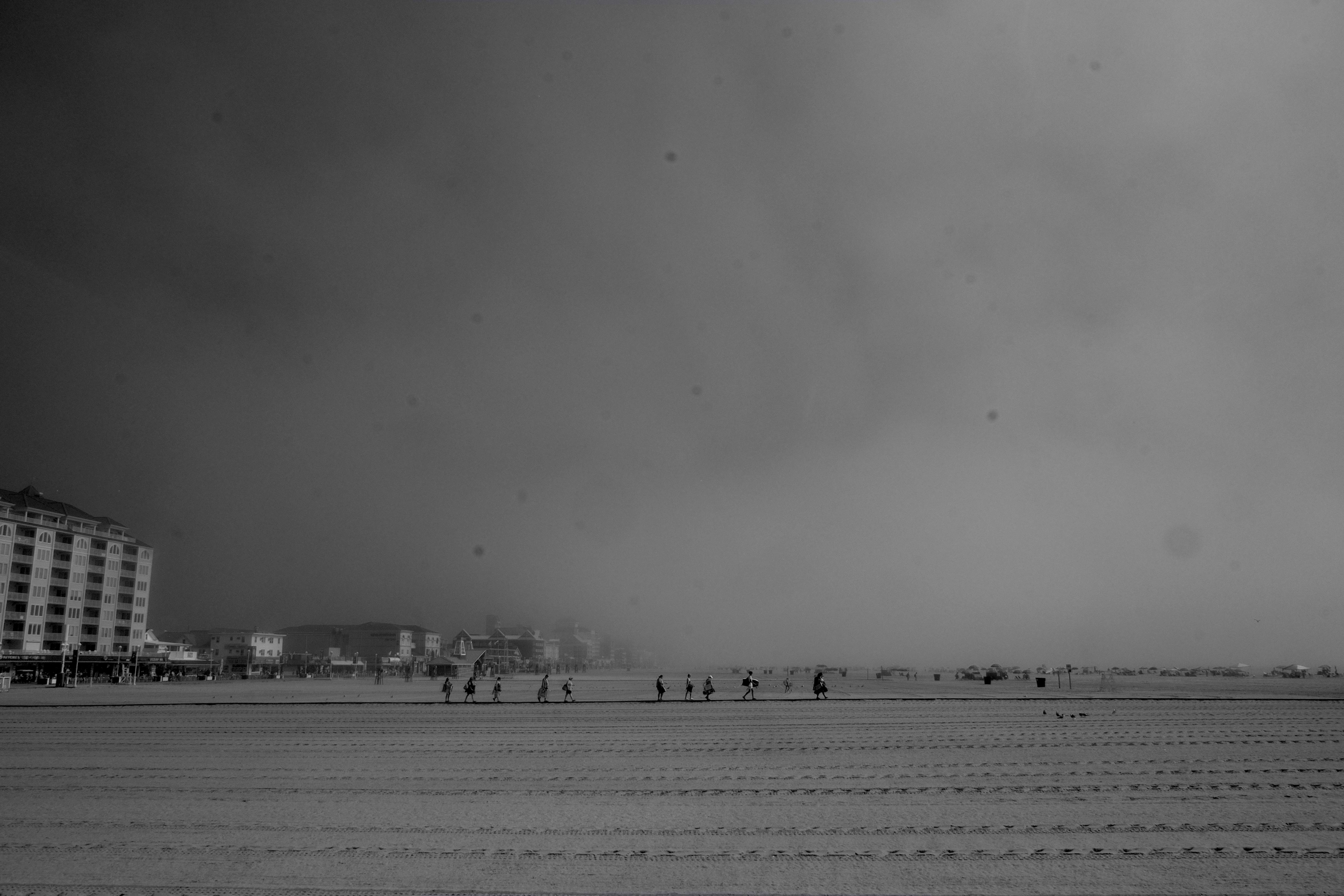 A black and white photo of a beach. People walk in the middle distance along a boardwalk, carrying beach supplies and boogie boards. Hotels and houses are visible in the distance, but fog obscures the sky.