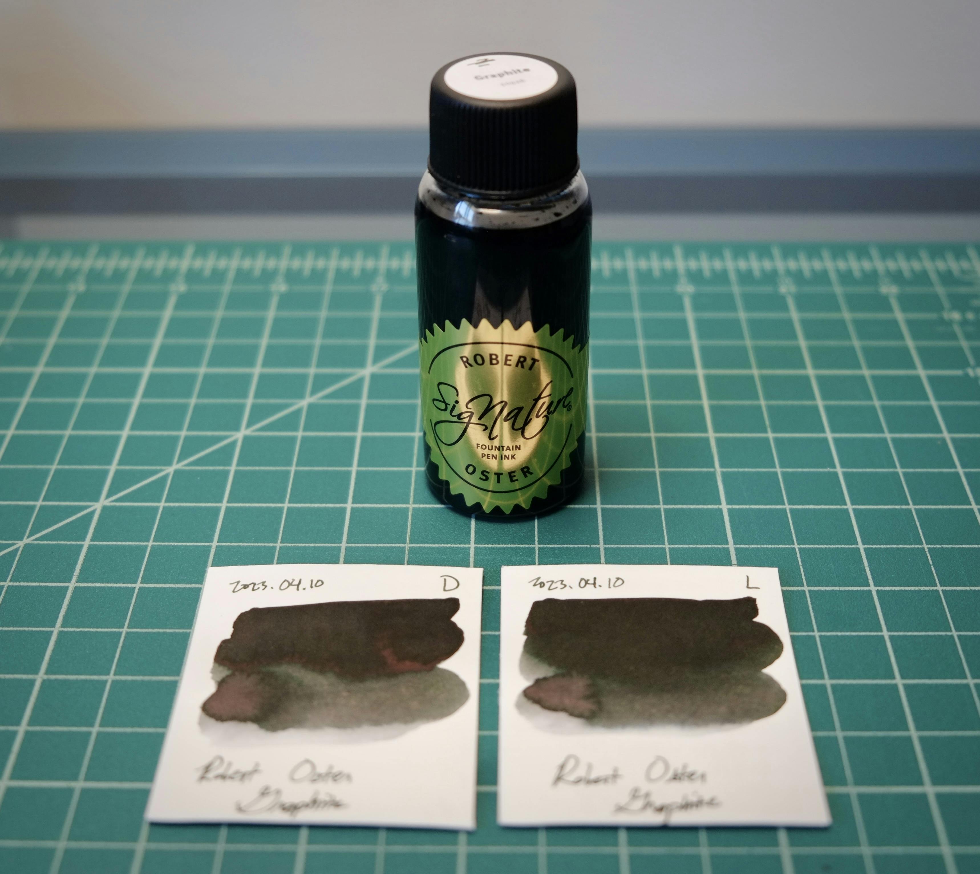 A bottle of Robert Oster Graphite ink on a desk, behind two color swatches