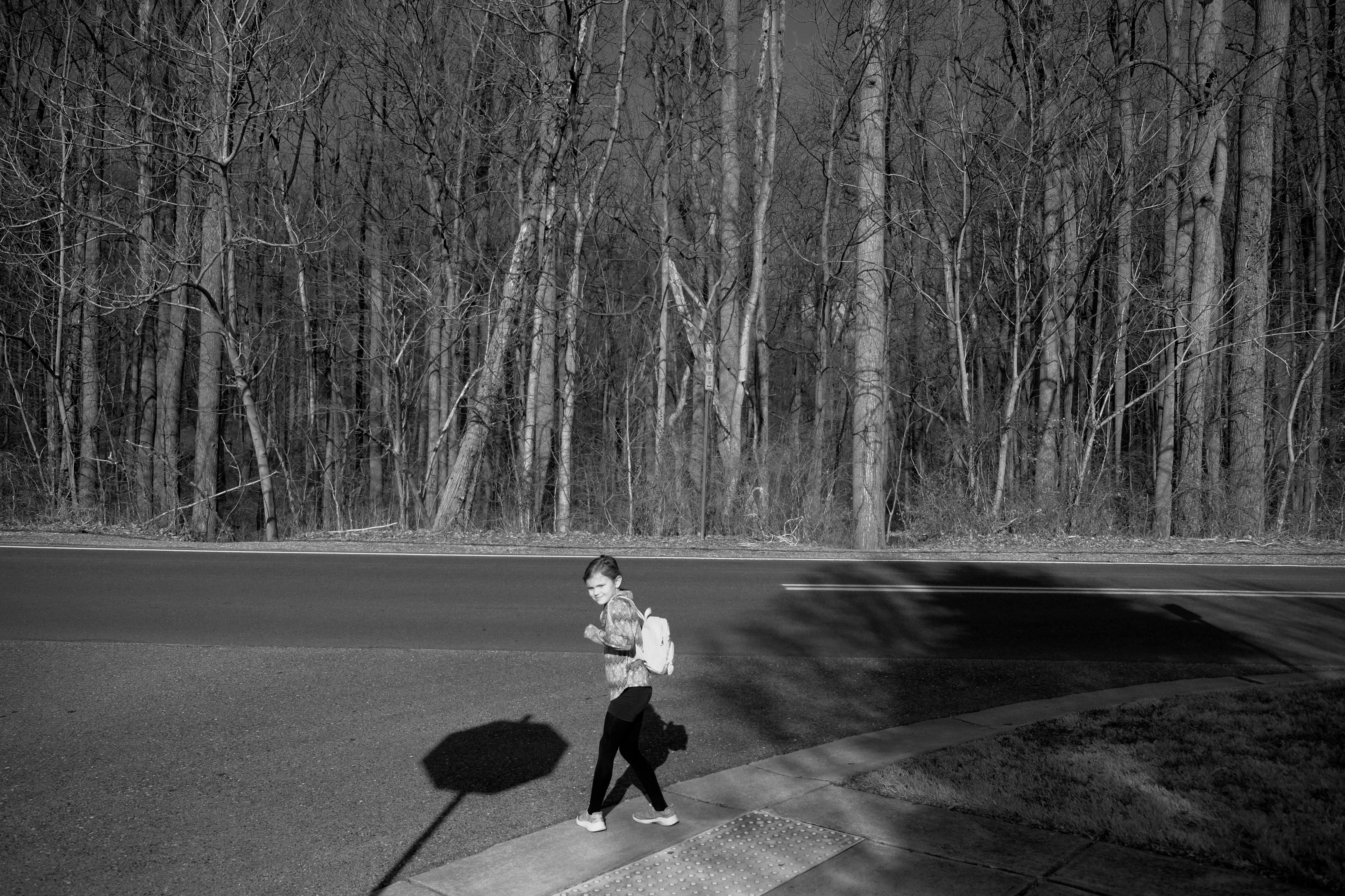 My daughter standing at a street corner, looking over her shoulder at the photographer. Across the street is a forest, with a trailhead just to the left.